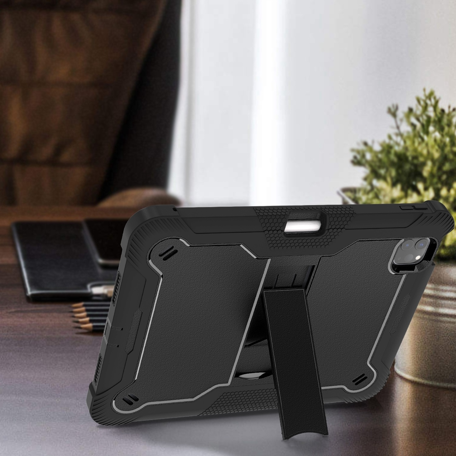 Tablet in schwarzer Hülle mit Ständer, auf einem Holztisch mit Bleistiften und Pflanze im Hintergrund.