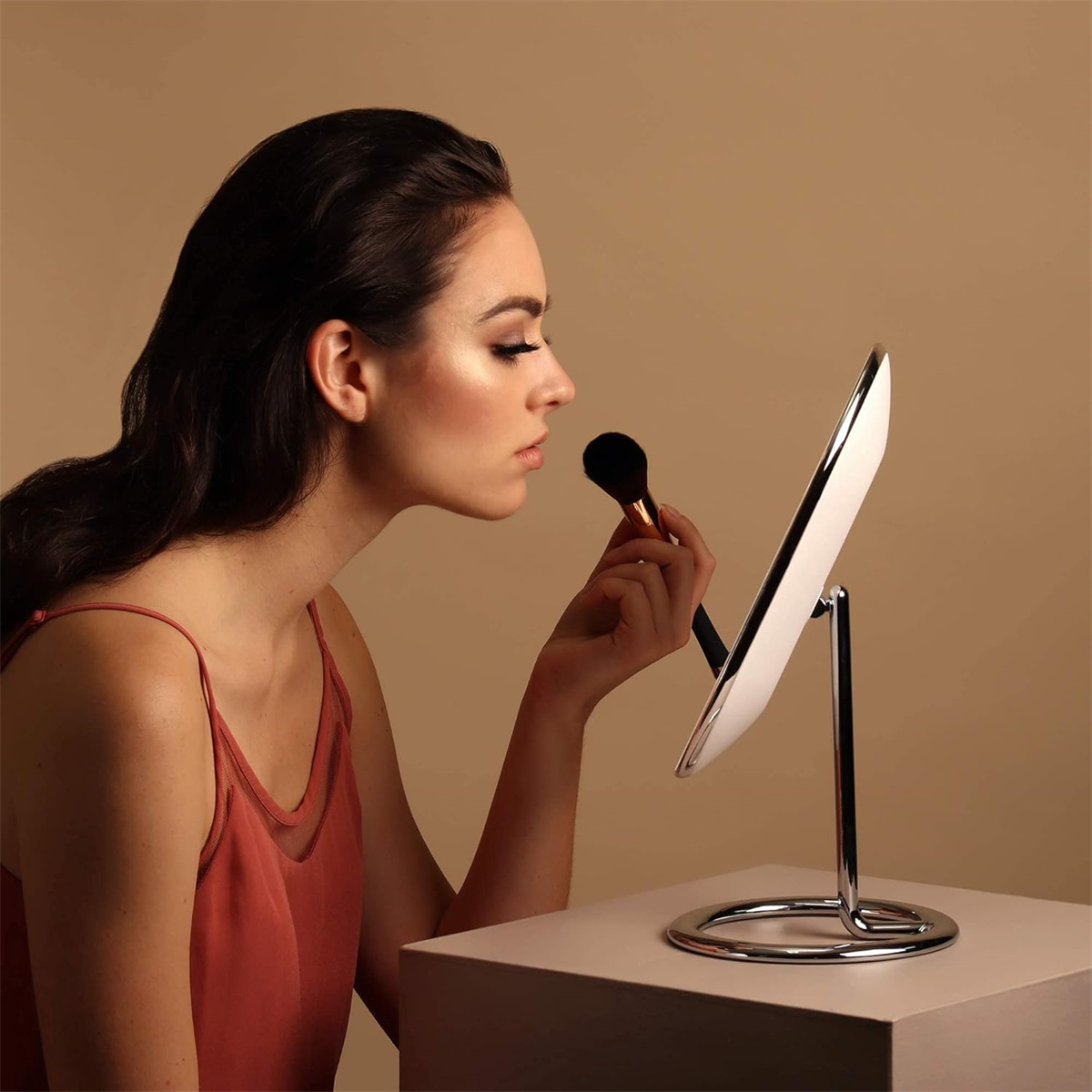 Eine Frau trägt Make-up mit einem Pinsel vor einem beleuchteten Spiegel auf.