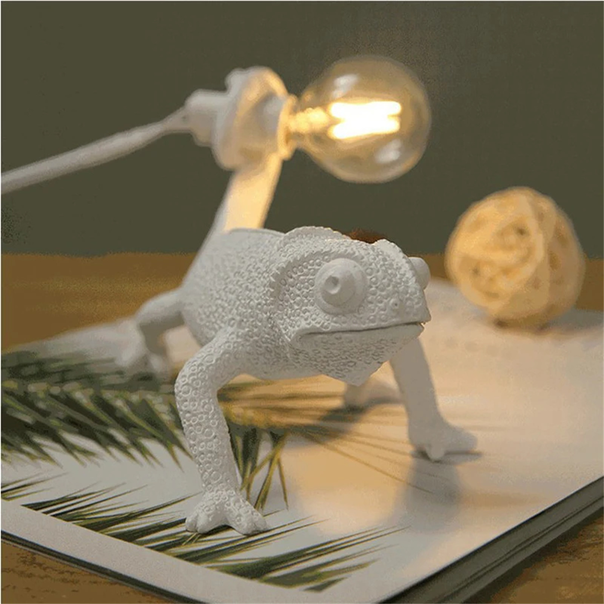 Weiße, chamäleonförmige Lampe mit Glühbirne, auf einem Buch platziert. Rechts ein Weidenball.