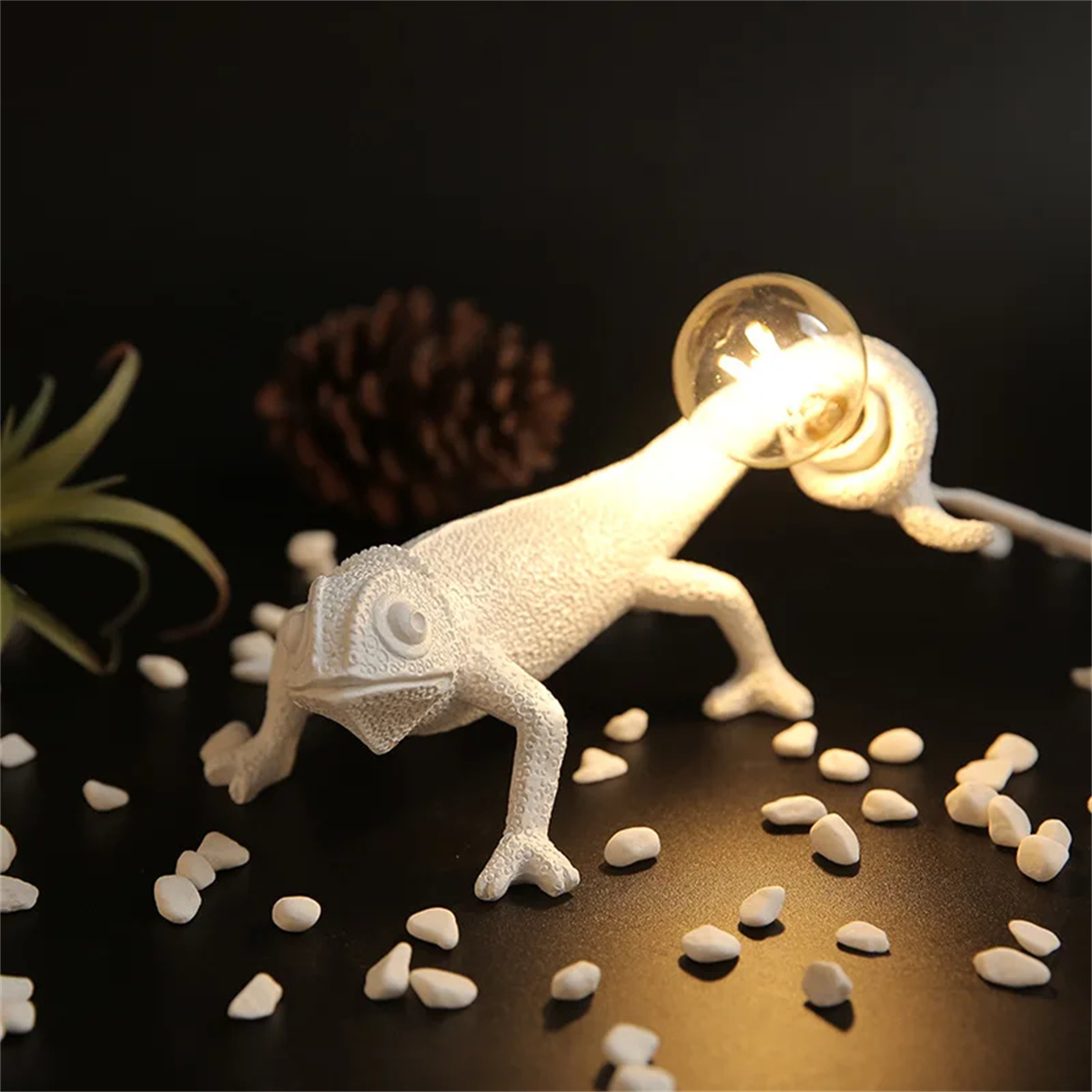 Eine weiße, chamäleonförmige Lampe mit leuchtender Glühbirne, auf schwarzem Hintergrund mit Kieselsteinen.