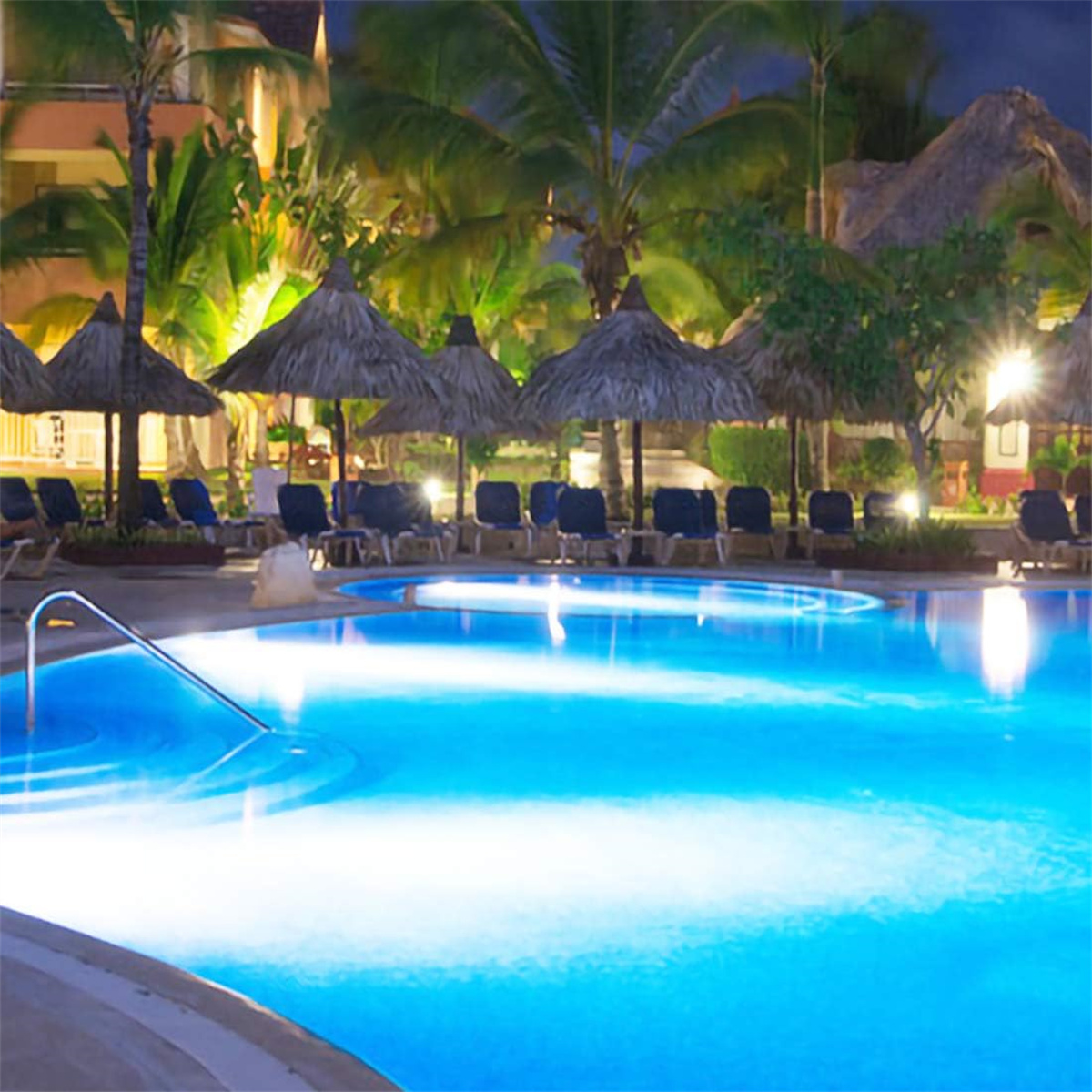 Eine Nachtszene eines Pools, Stühlen und Palmen, mit einem Resort im Hintergrund.