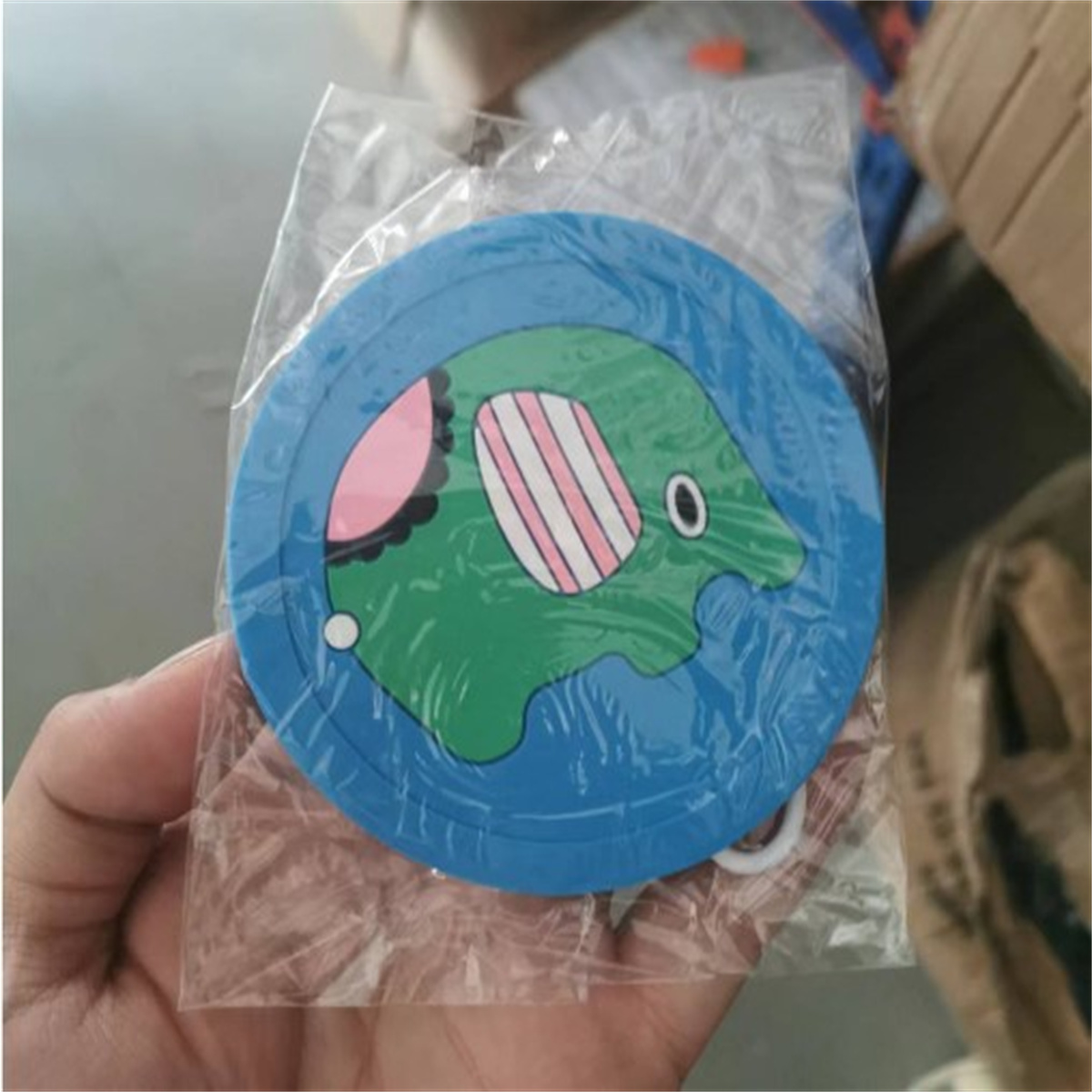 Ein blau-grüner, elefantenförmiger Untersetzer wird in einer Hand gehalten, in einer durchsichtigen Plastikverpackung.