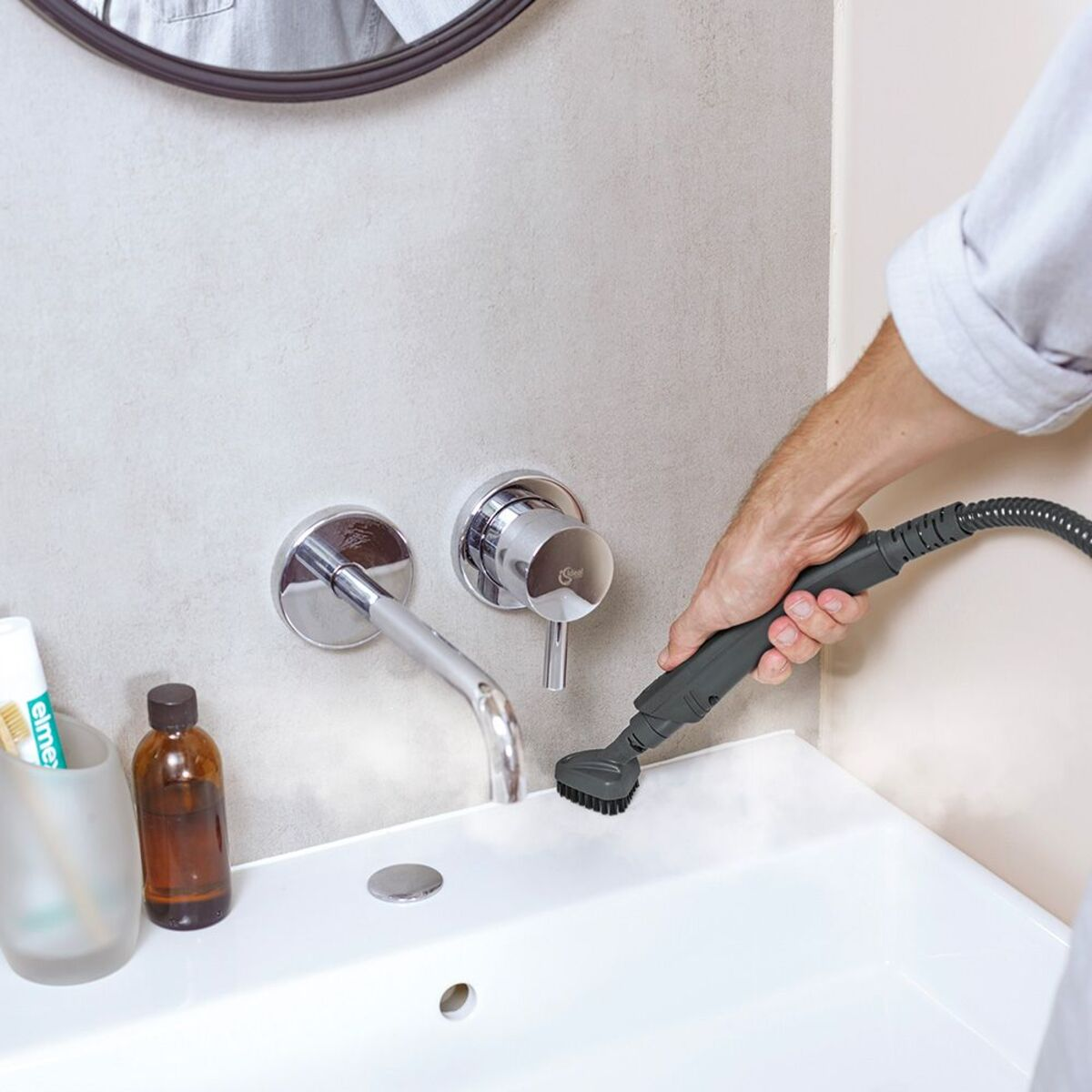 Osoba czyszcząca kran w zlewie za pomocą parownicy. Zlew jest biały, a ściana szara.