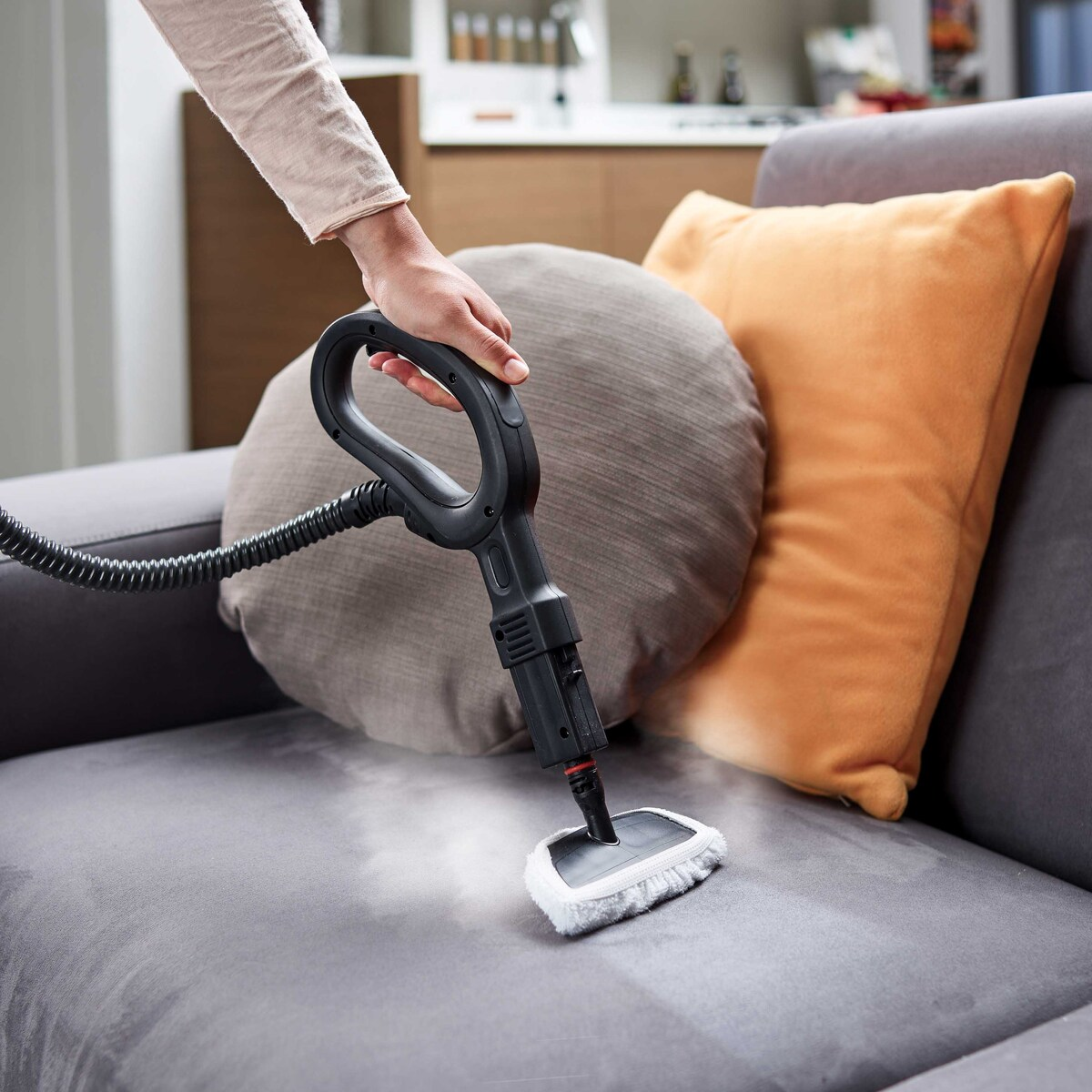 Una mano usando una limpiadora a vapor en un sofá gris. Hay almohadas en el fondo.