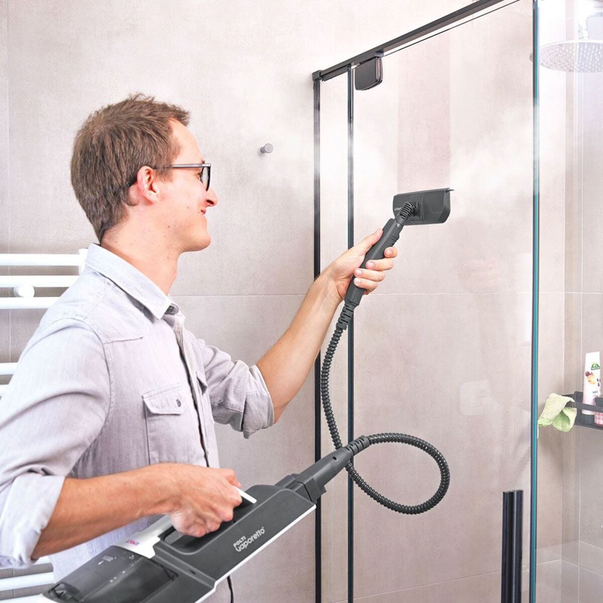 Mężczyzna czyszczący szybę prysznicową za pomocą parownicy. Ma okulary i szarą koszulę.