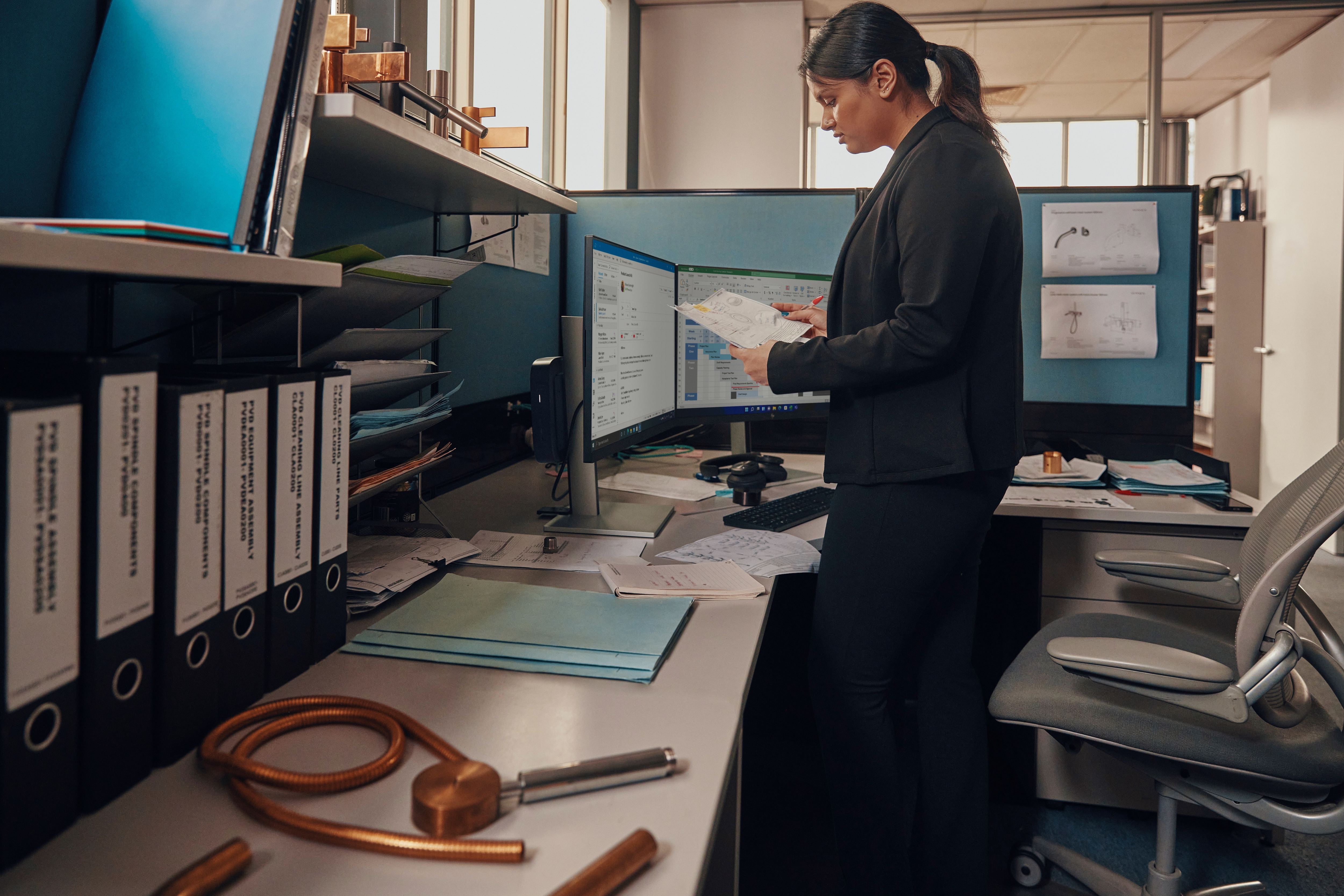 Donna in ufficio, guarda gli schermi dei computer. Documenti ed equipaggiamento sulla scrivania. Luce naturale.