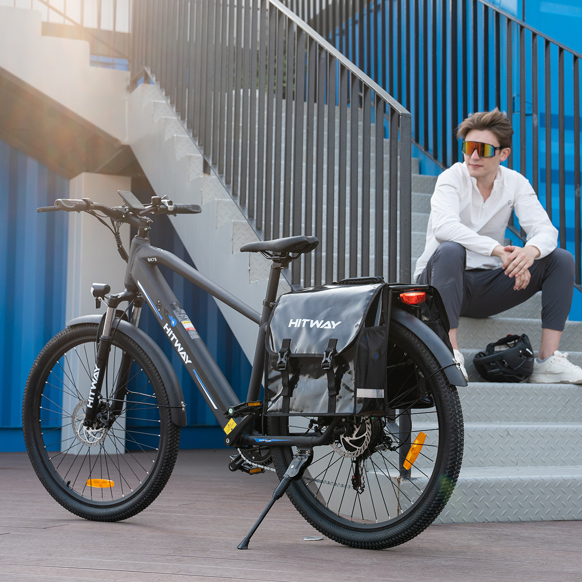 Mann sitzt auf Treppe neben geparktem schwarzen HITWAY E-Bike mit Zubehör. Blauer Hintergrund.