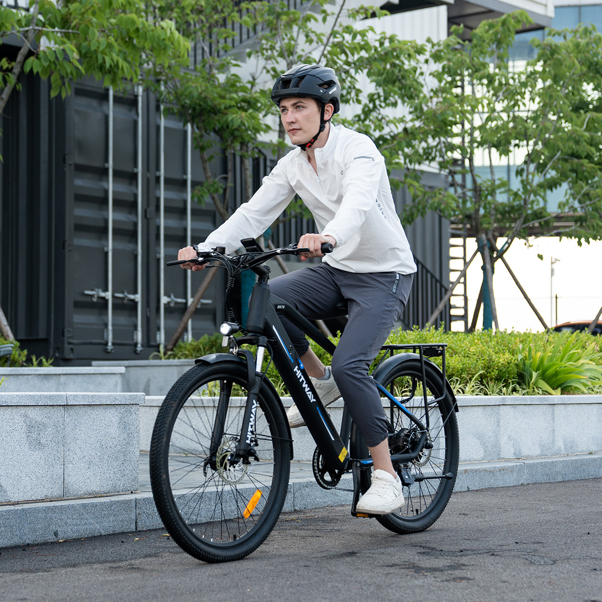 Ein Mann mit Helm fährt auf einem asphaltierten Weg ein schwarzes Elektrofahrrad in der Nähe eines Gebäudes.