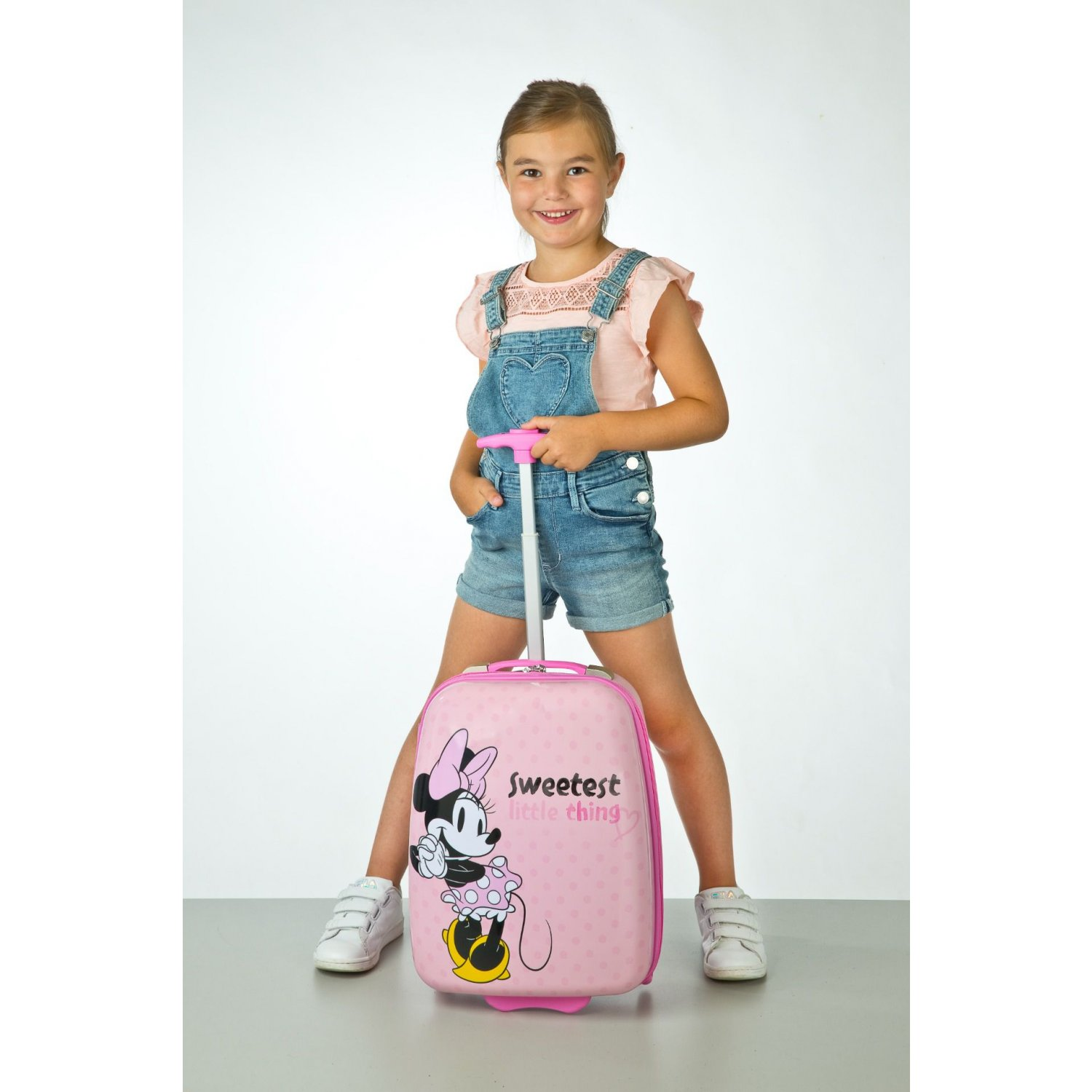 Mädchen mit einem rosa Minnie-Maus-Koffer auf weißem Hintergrund. Sie lächelt und trägt Jeansshorts.