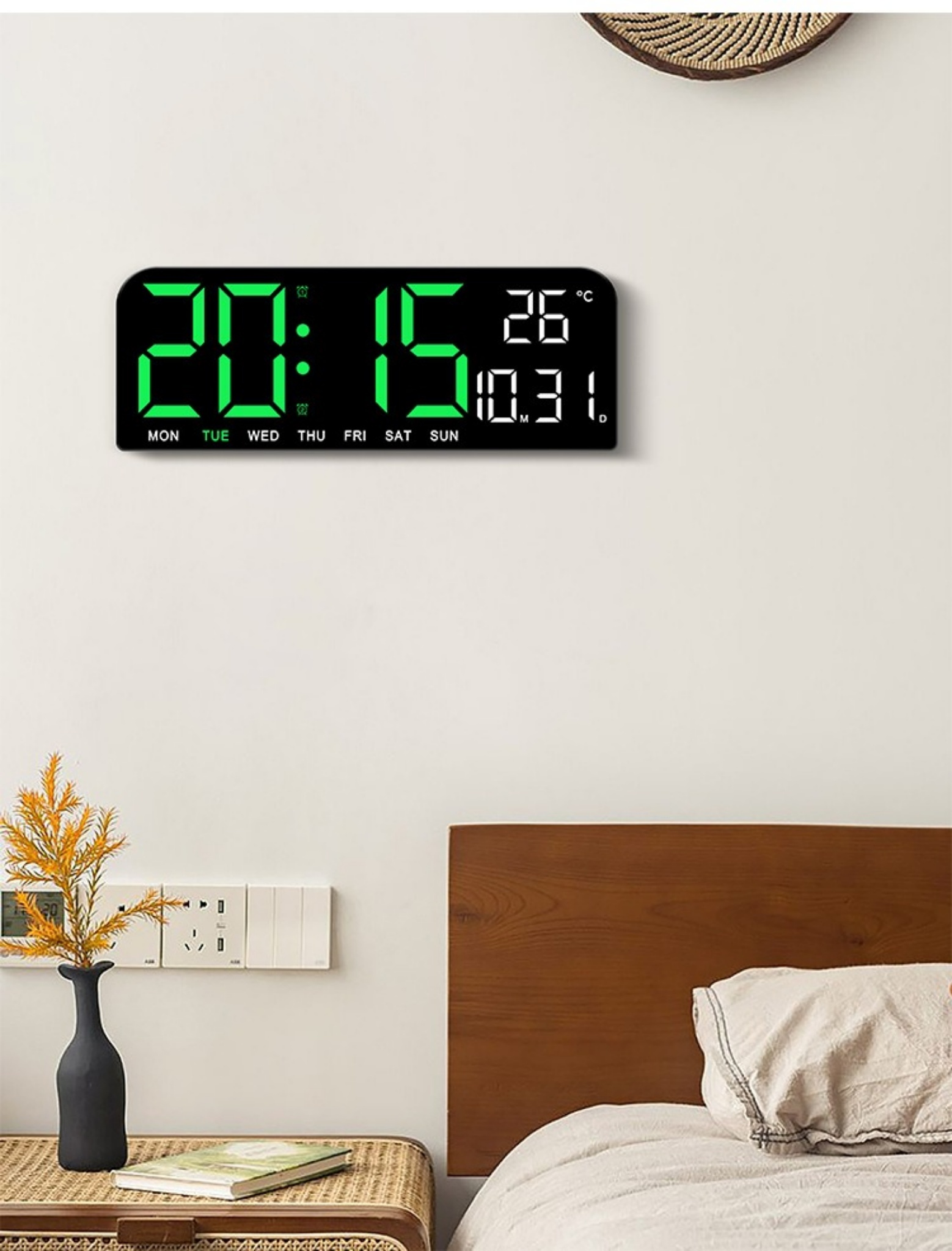 Digitaluhr an der Wand, zeigt Zeit und Datum an. Im Hintergrund ein Schlafzimmer mit Bett und Wand.