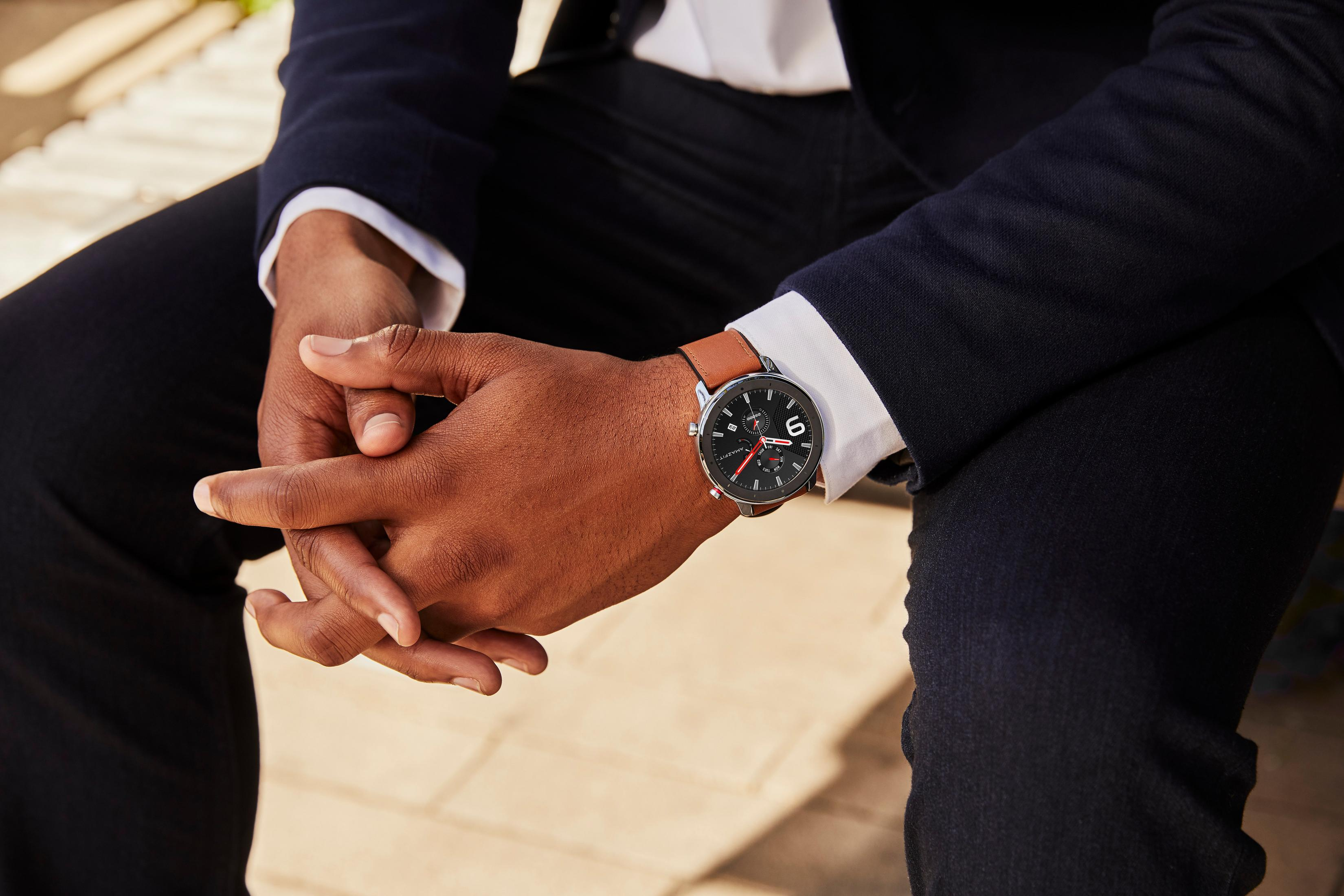 Person trägt eine Uhr mit schwarzem Zifferblatt und braunem Lederarmband. Hände gefaltet.