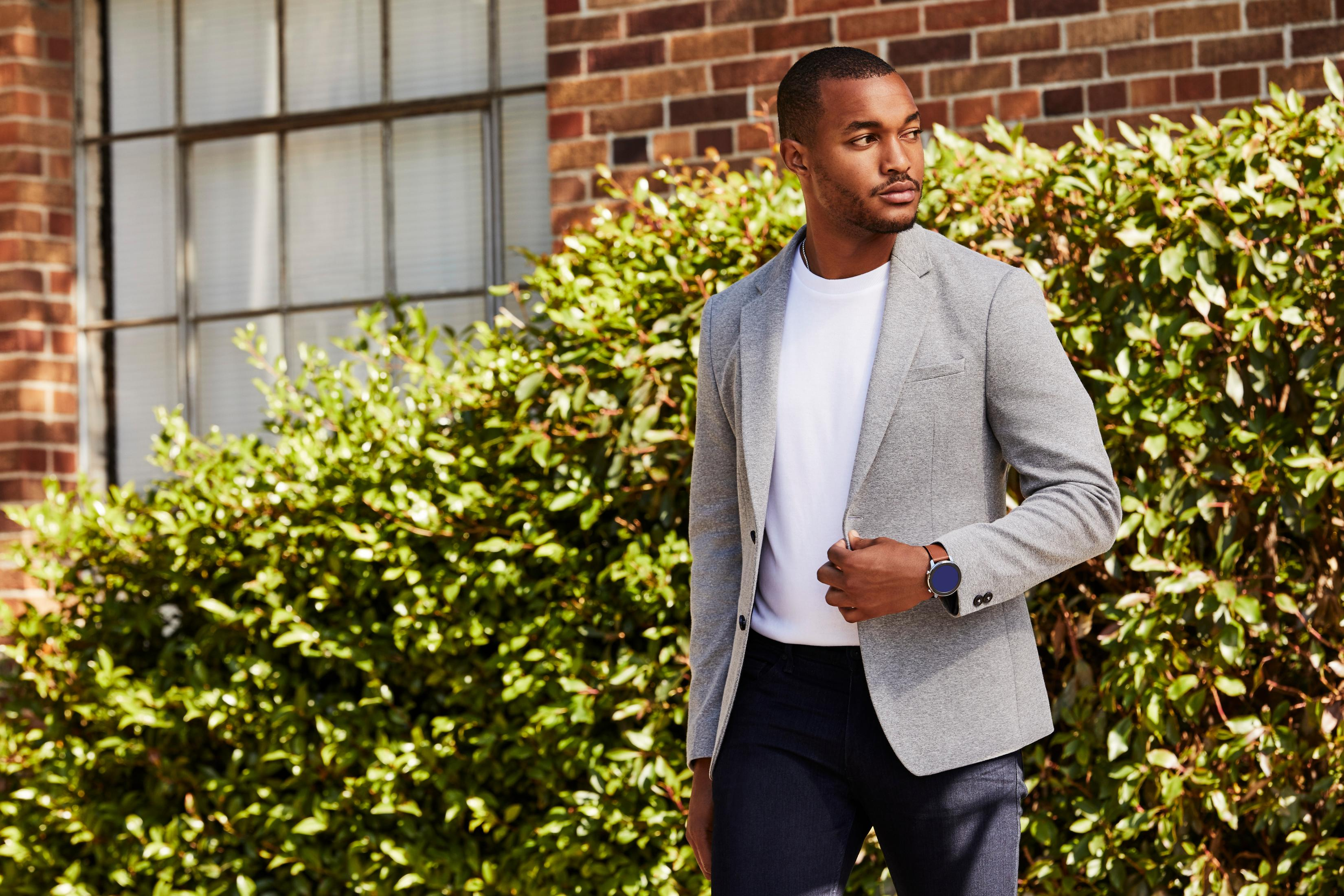 Mann in grauem Blazer, weißem Hemd und dunkler Hose neben grünem Busch.