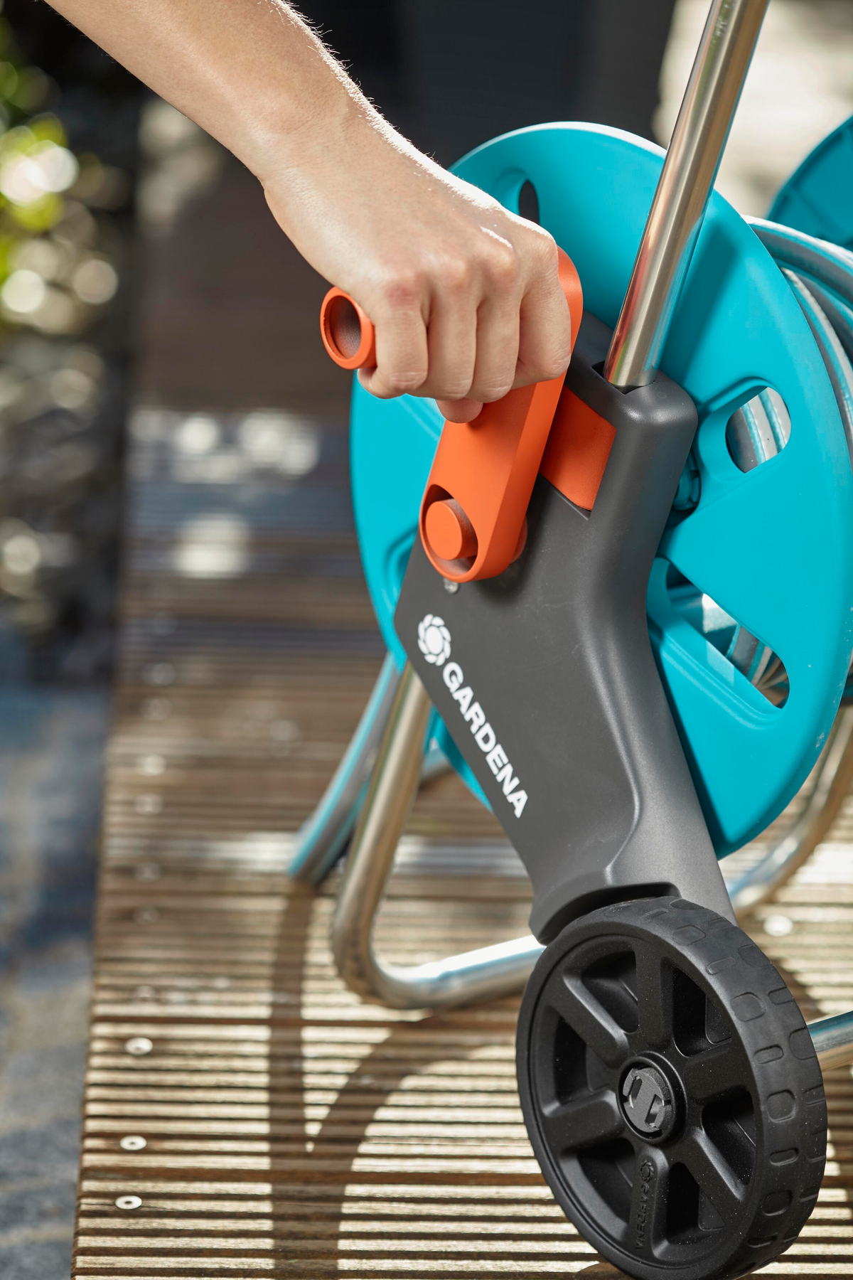 Hand dreht die orangefarbene Kurbel einer blauen Schlauchtrommel. Räder sind schwarz. Holzboden im Hintergrund.