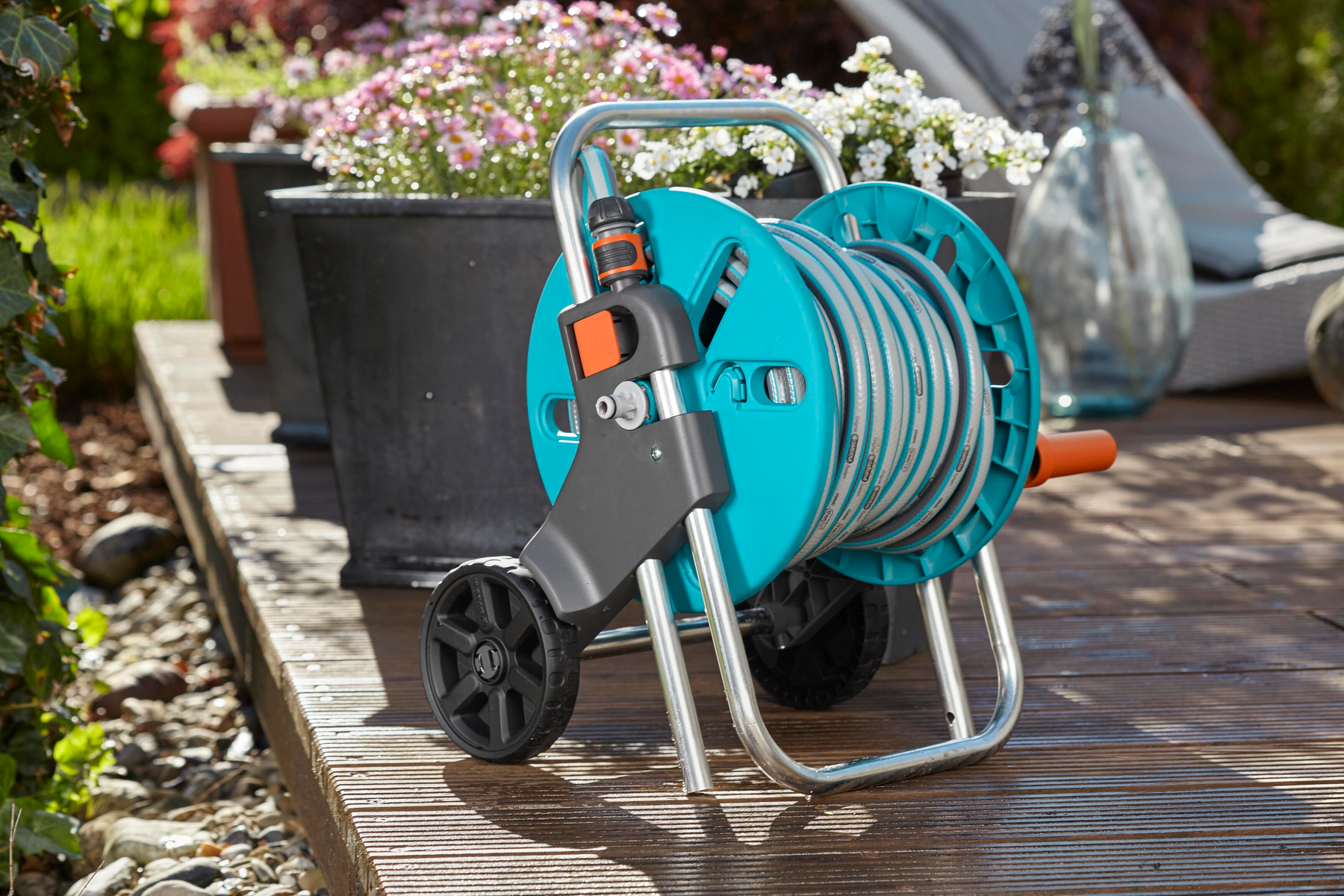 Eine türkisfarbene Schlauchtrommel auf Rädern mit Gartenschlauch. Dahinter Töpfe und Blumen.