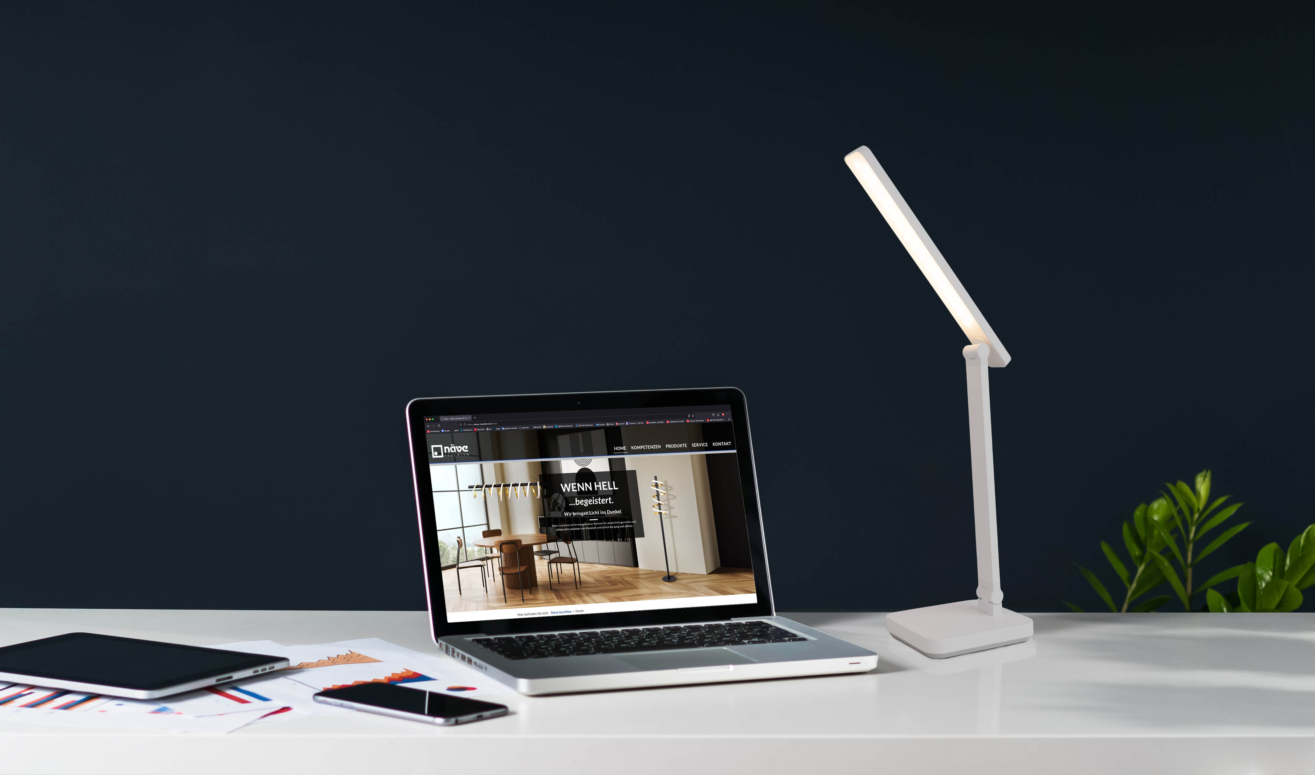 Ein Laptop, ein Tablet und eine Lampe auf einem weißen Schreibtisch vor einer dunkelblauen Wand.