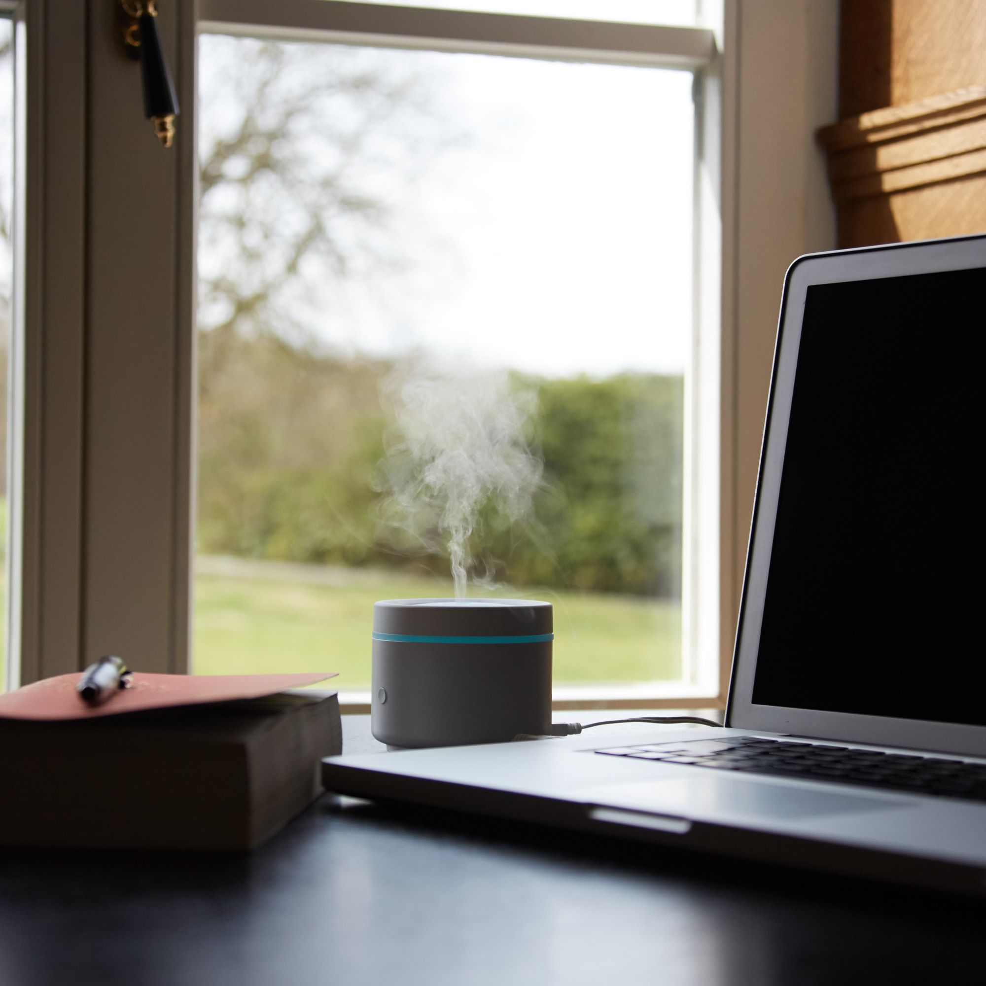 Humidifier with vapor, a laptop, and a book on a desk near a window.