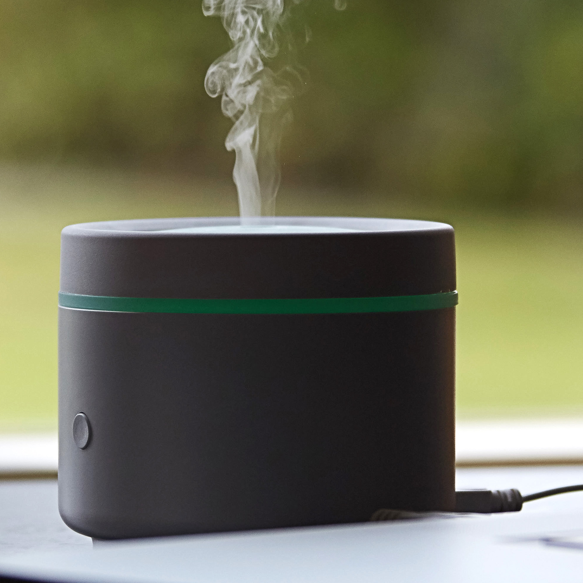 A dark gray essential oil diffuser with a green stripe, releasing steam. A button and cord are visible.