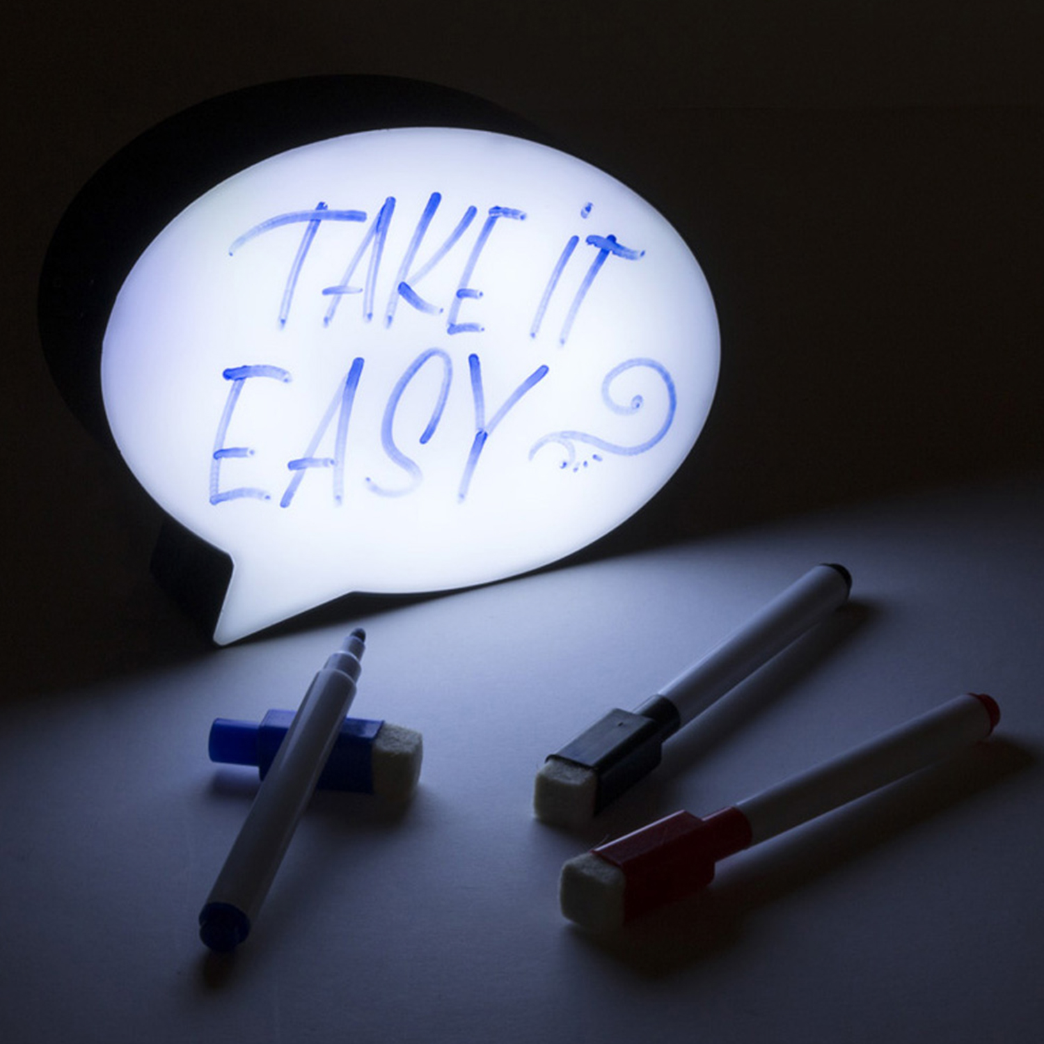 Beleuchtete Sprechblase mit der Aufschrift 'TAKE IT EASY'. Drei Stifte und ein Radiergummi auf einer Oberfläche. Dunkler Hintergrund.