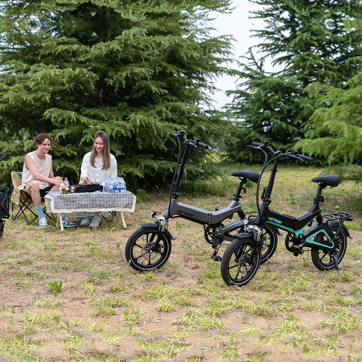 Zwei schwarze und graue HITWAY-Fahrräder auf Gras. Zwei Personen sitzen im Hintergrund an einem Tisch.