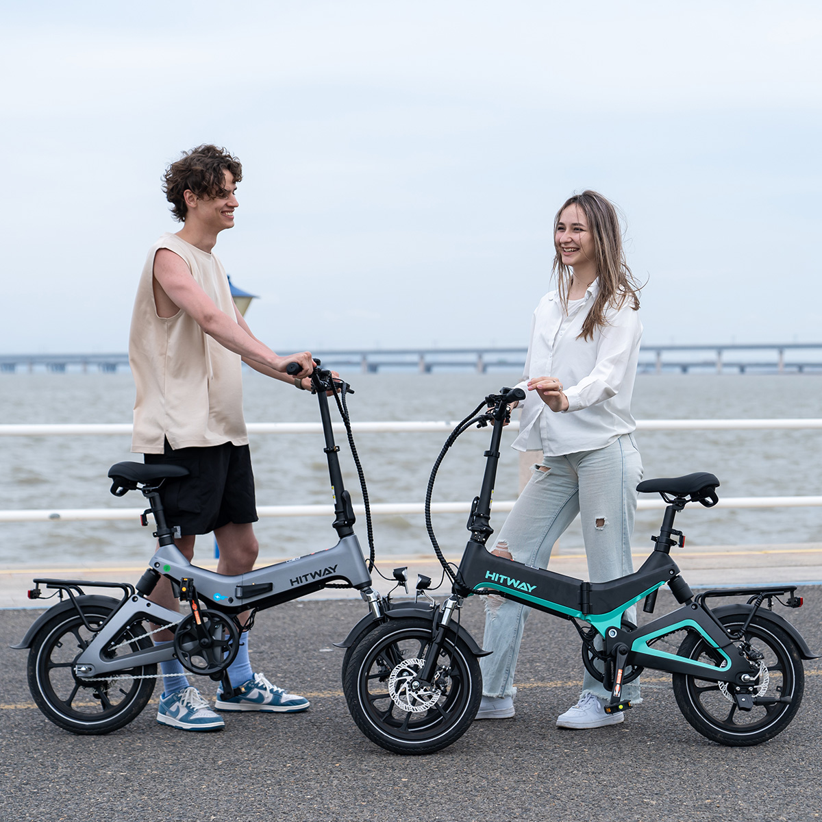 Zwei Personen mit E-Bikes stehen auf einem Weg. Fahrräder sind grau und türkis. Hintergrund: Wasser und Brücke.