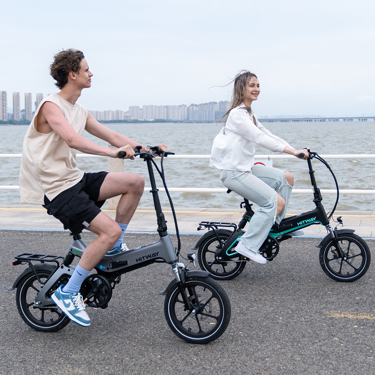 Zwei Personen fahren nebeneinander auf einem grauen und schwarzen Elektrofahrrad auf einem gepflasterten Weg in Wassernähe.