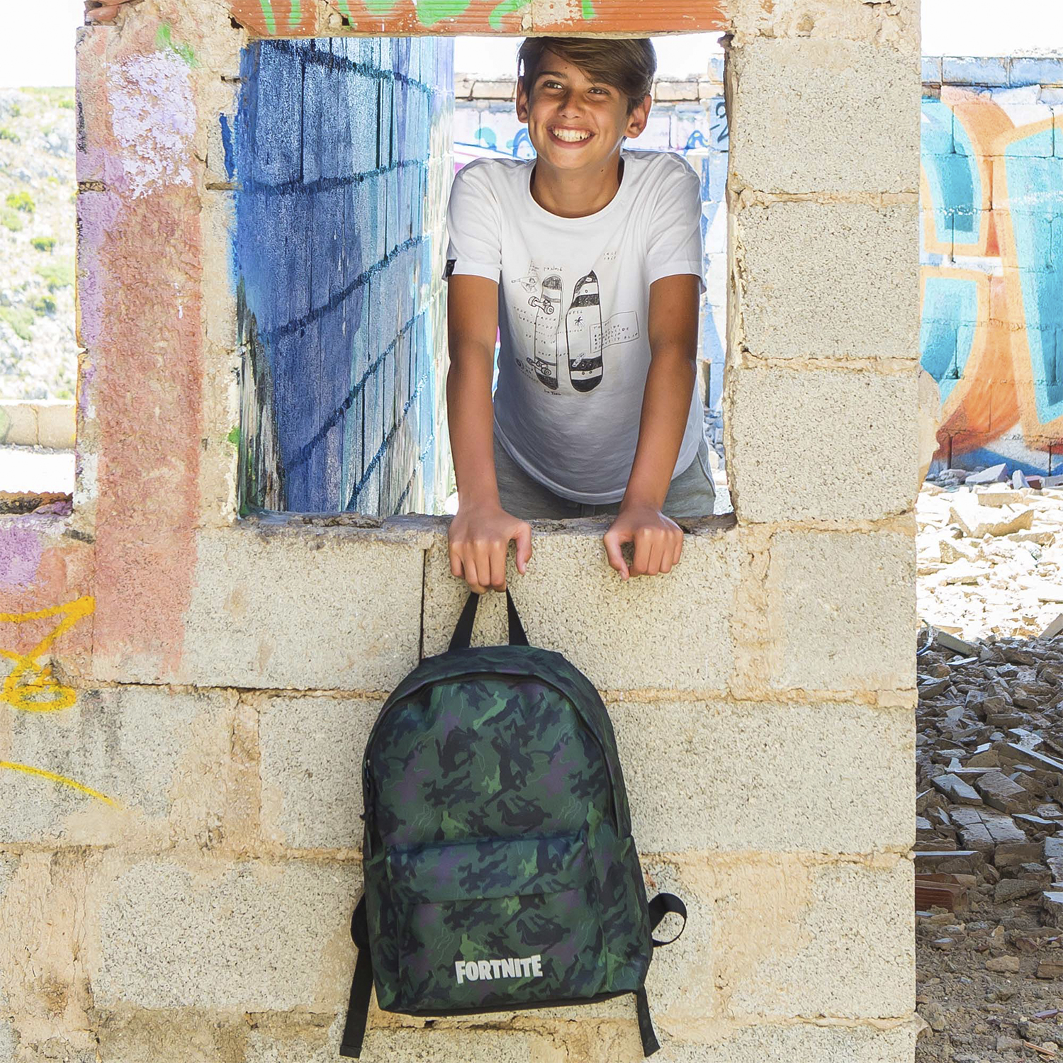 Un ragazzo sorride da una finestra di cemento, con uno zaino Fortnite in un ambiente graffiti.