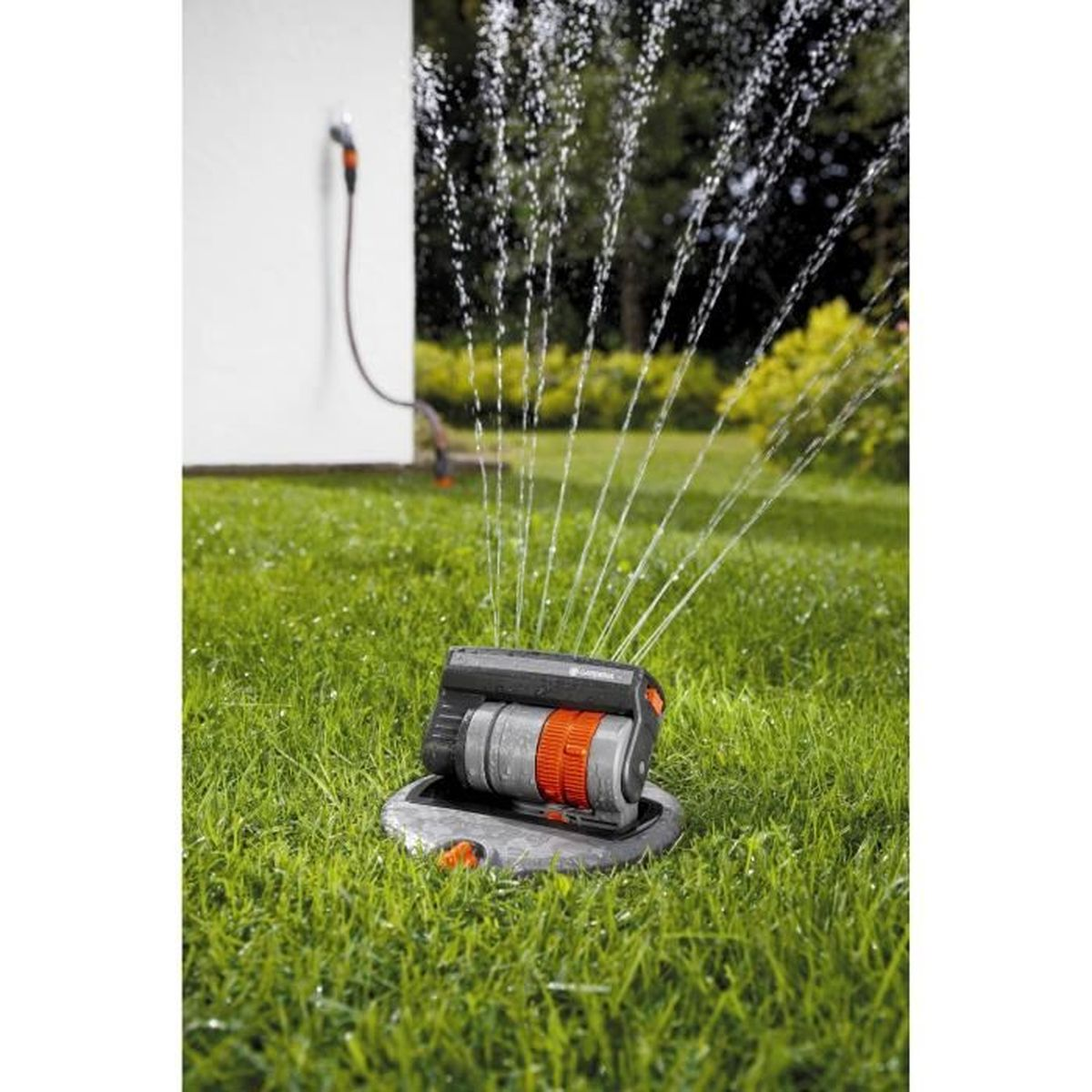 Rasensprenger sprüht Wasser auf grünes Gras, mit weißer Wand und Bäumen im Hintergrund.
