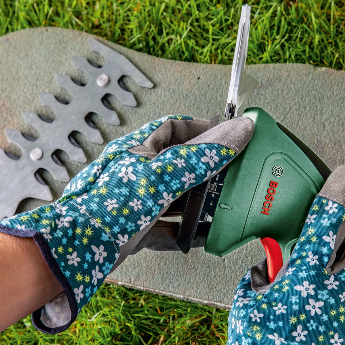 Hände in Blumenhandschuhen verwenden eine grüne Bosch Heckenschere. Klingen sind sichtbar. Grüner Gras Hintergrund.