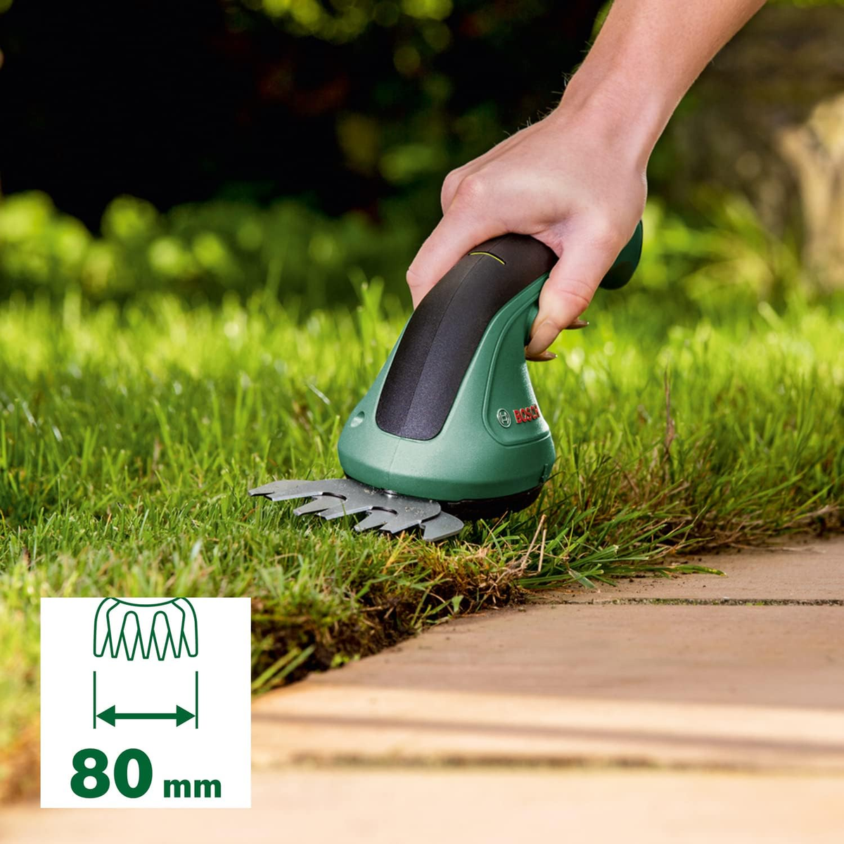 Grüne und schwarze elektrische Grasschere schneidet Gras. Das Werkzeug wird von einer Hand gehalten. 80mm Messung.