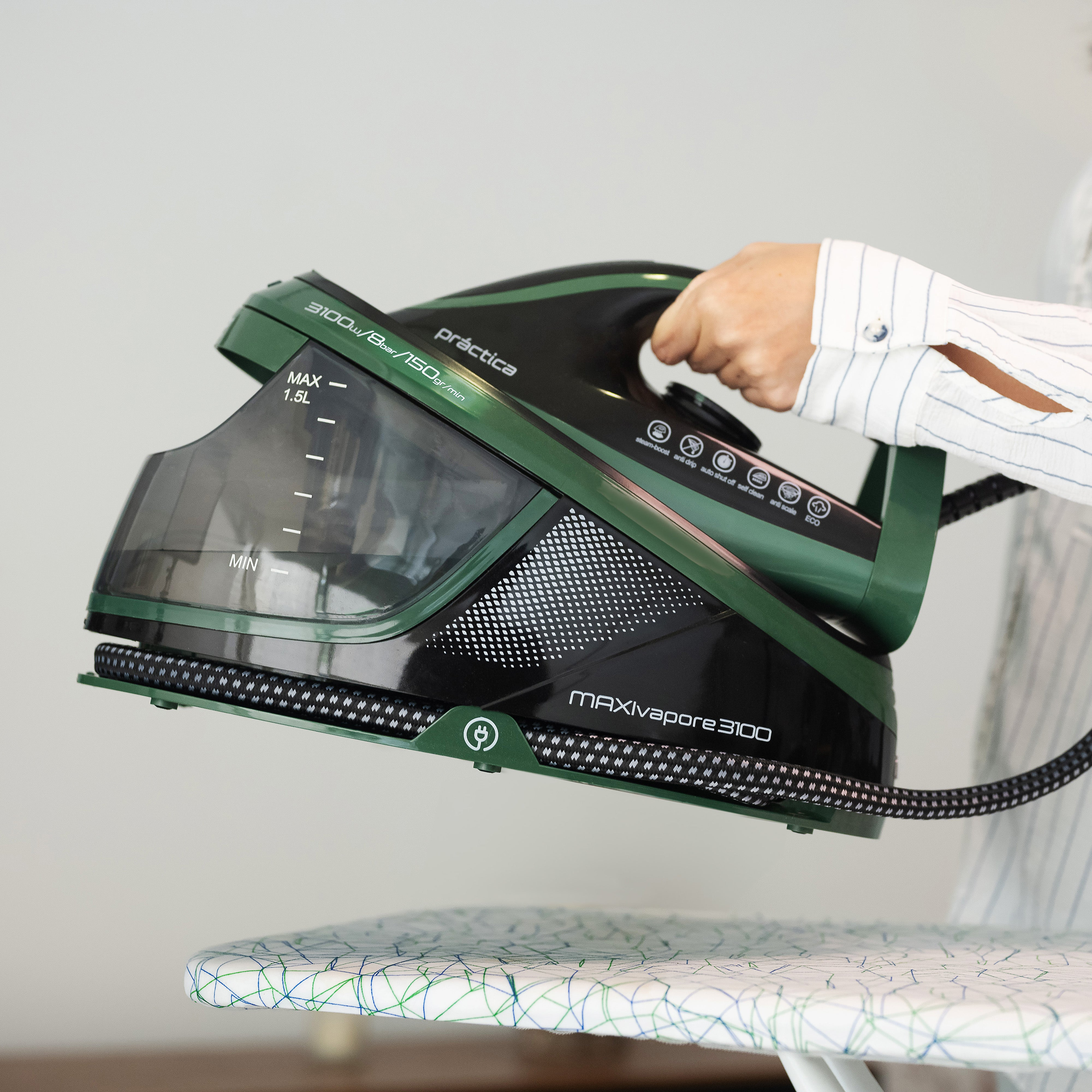 Persona sosteniendo una plancha verde y negra sobre una tabla de planchar. La plancha está echando vapor.