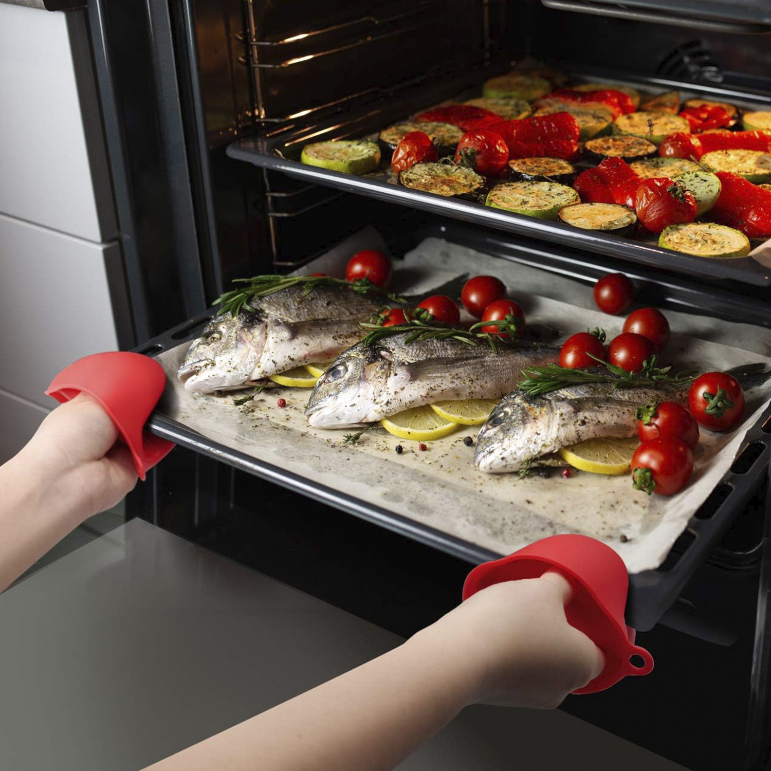 Una persona sosteniendo una bandeja de pescado horneado con tomates y limones en el horno.