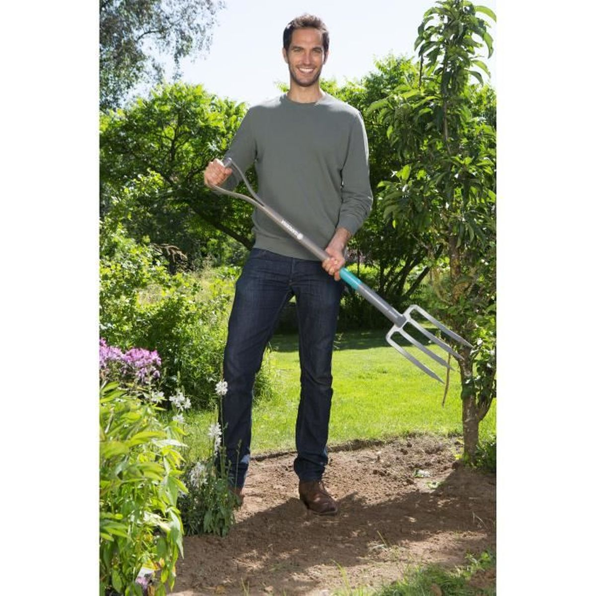 Un uomo che tiene una forca da giardino in un giardino, sorridendo. Indossa jeans e una maglia verde a maniche lunghe.
