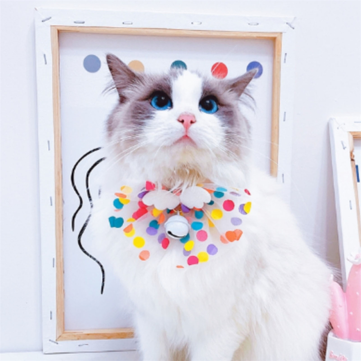 Eine flauschige Katze mit blauen Augen trägt ein buntes Halsband, sitzt vor einer Zeichnung.