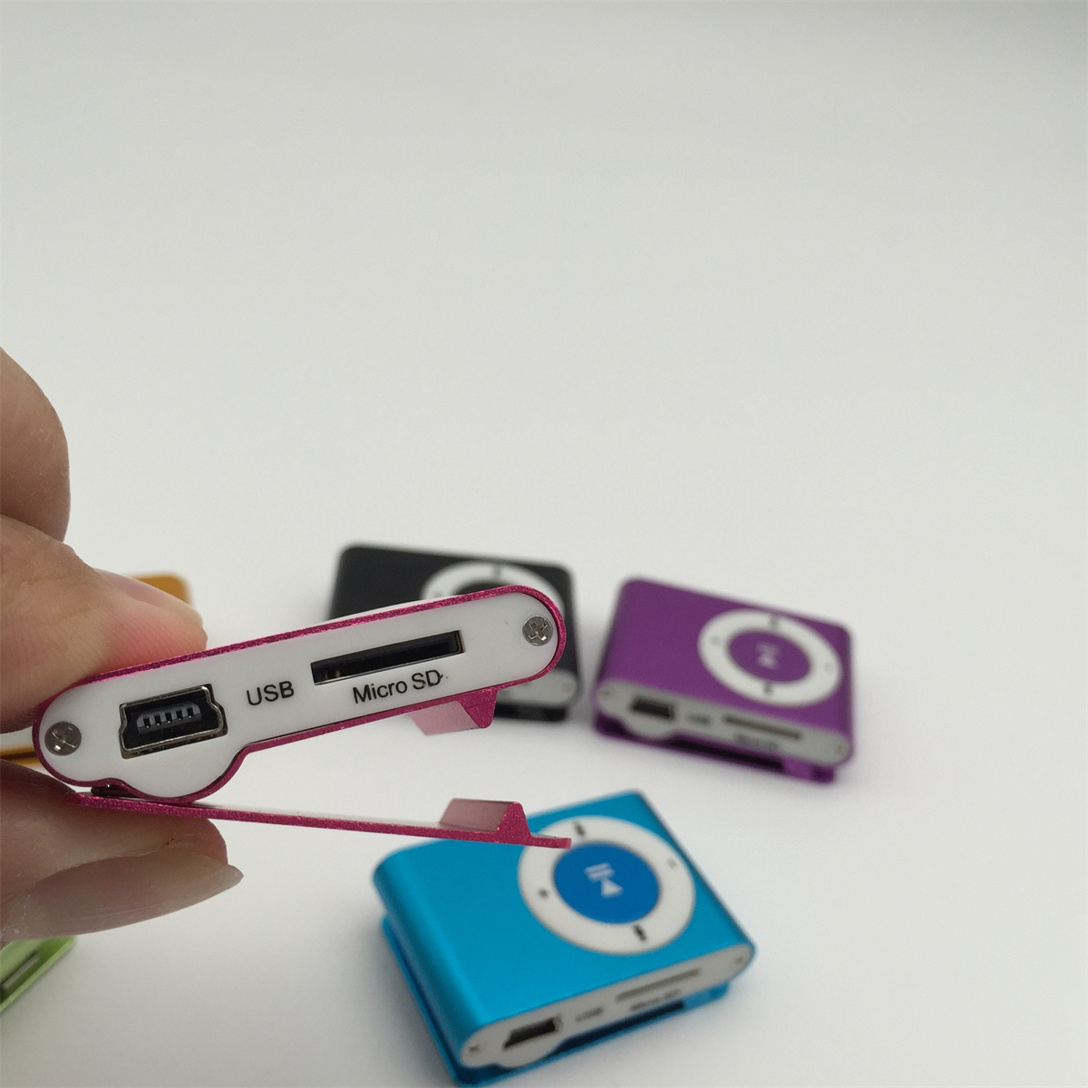 Eine Hand hält einen pinkfarbenen MP3-Player. Andere MP3-Player in Schwarz, Lila und Blau liegen auf dem Tisch.