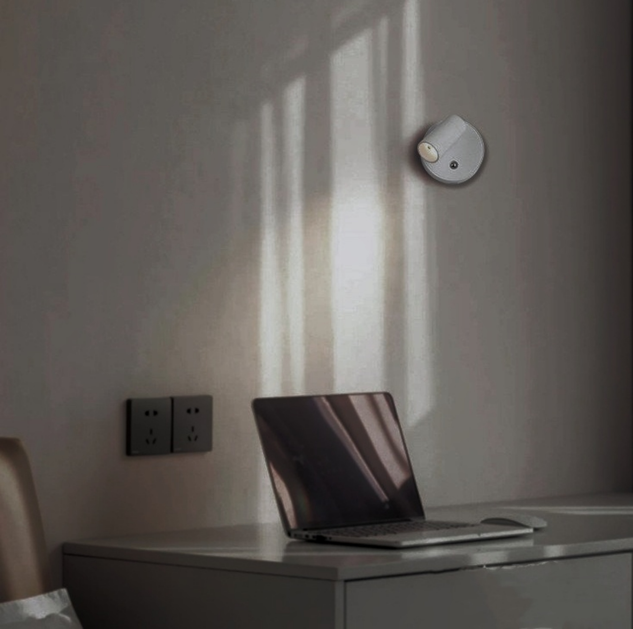 Ein Laptop und ein kleines Licht neben einer Steckdose an einer grauen Wand. Die Wand wird teilweise von Sonnenlicht beleuchtet.