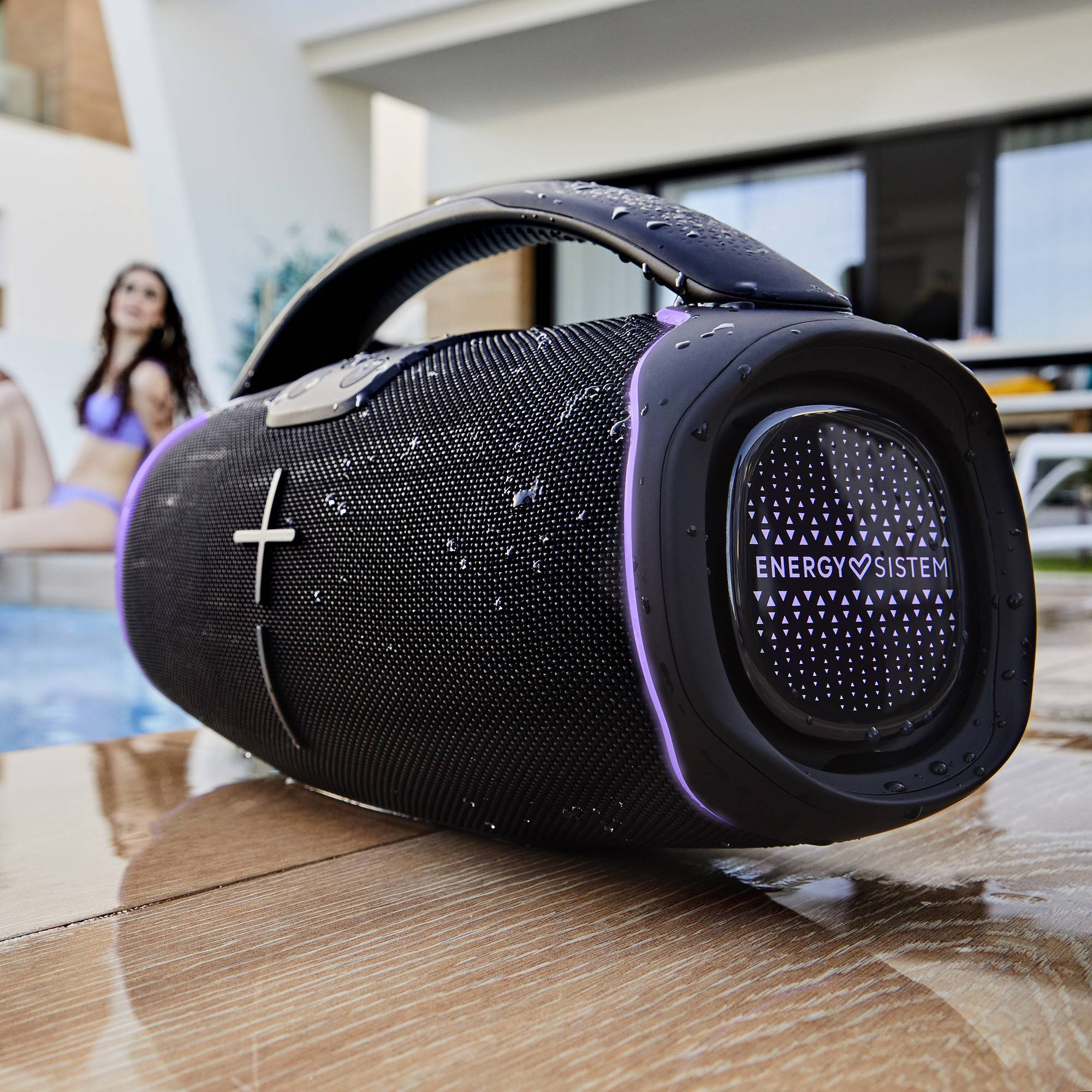 Altoparlante nero con goccioline d'acqua, maniglia, finiture viola e una donna sullo sfondo.