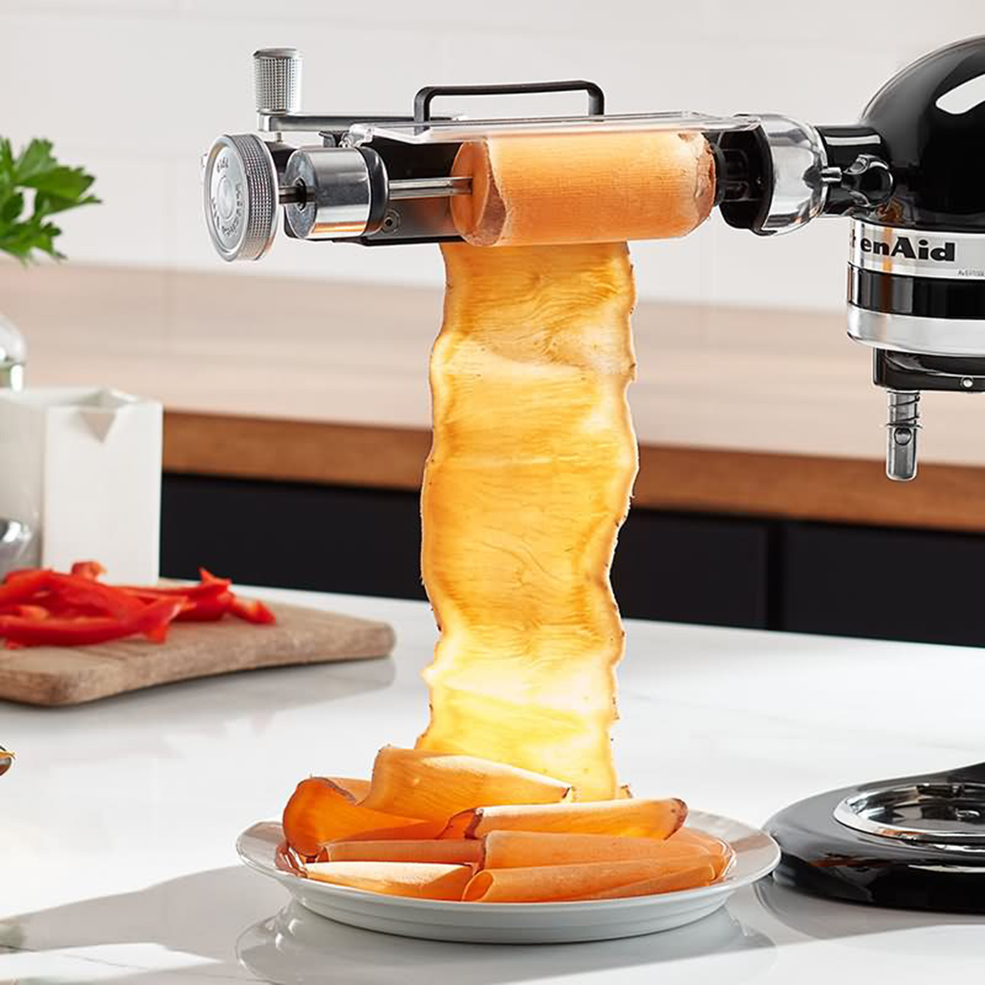 KitchenAid-Gerät mit Gemüse-Spiralschneider-Aufsatz. Zeigt, wie Karotten spiralförmig geschnitten werden und auf einen Teller fallen.