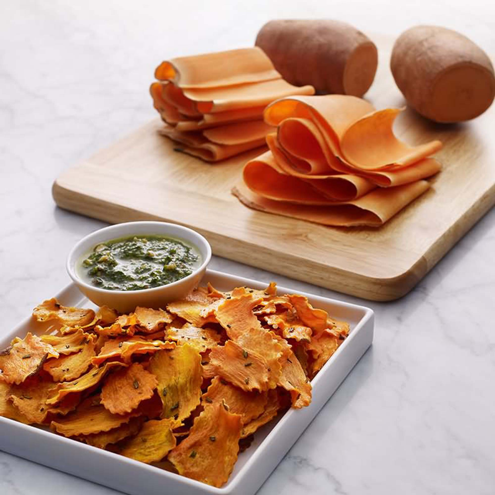 Nahaufnahme von Süßkartoffelchips und Dip auf einem Holzbrett. Zwei Kartoffeln sind im Hintergrund. Weiße Marmoroberfläche.