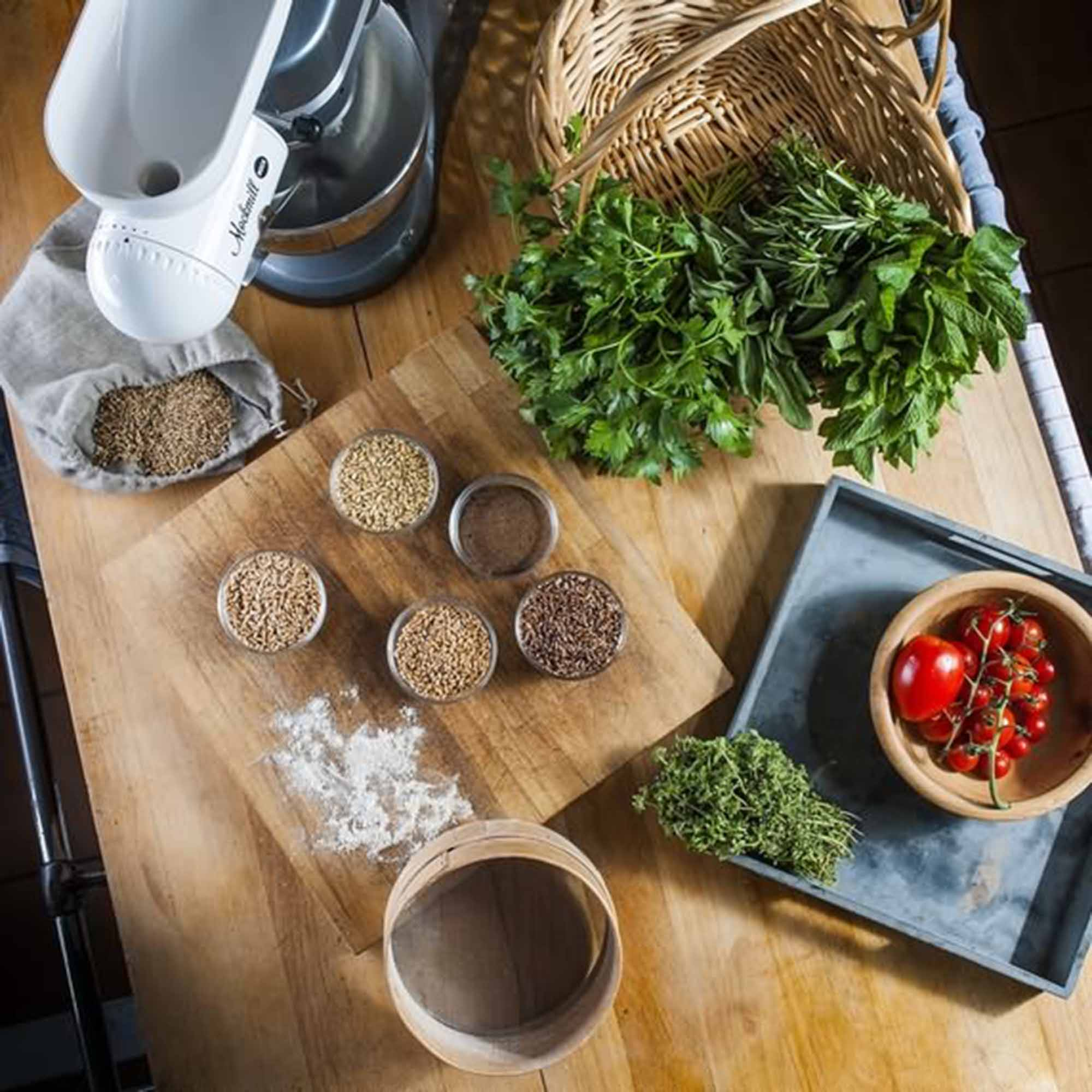 Scena kuchenna ze składnikami na drewnianej desce. Zawiera zioła, pomidory i młyn do żywności.