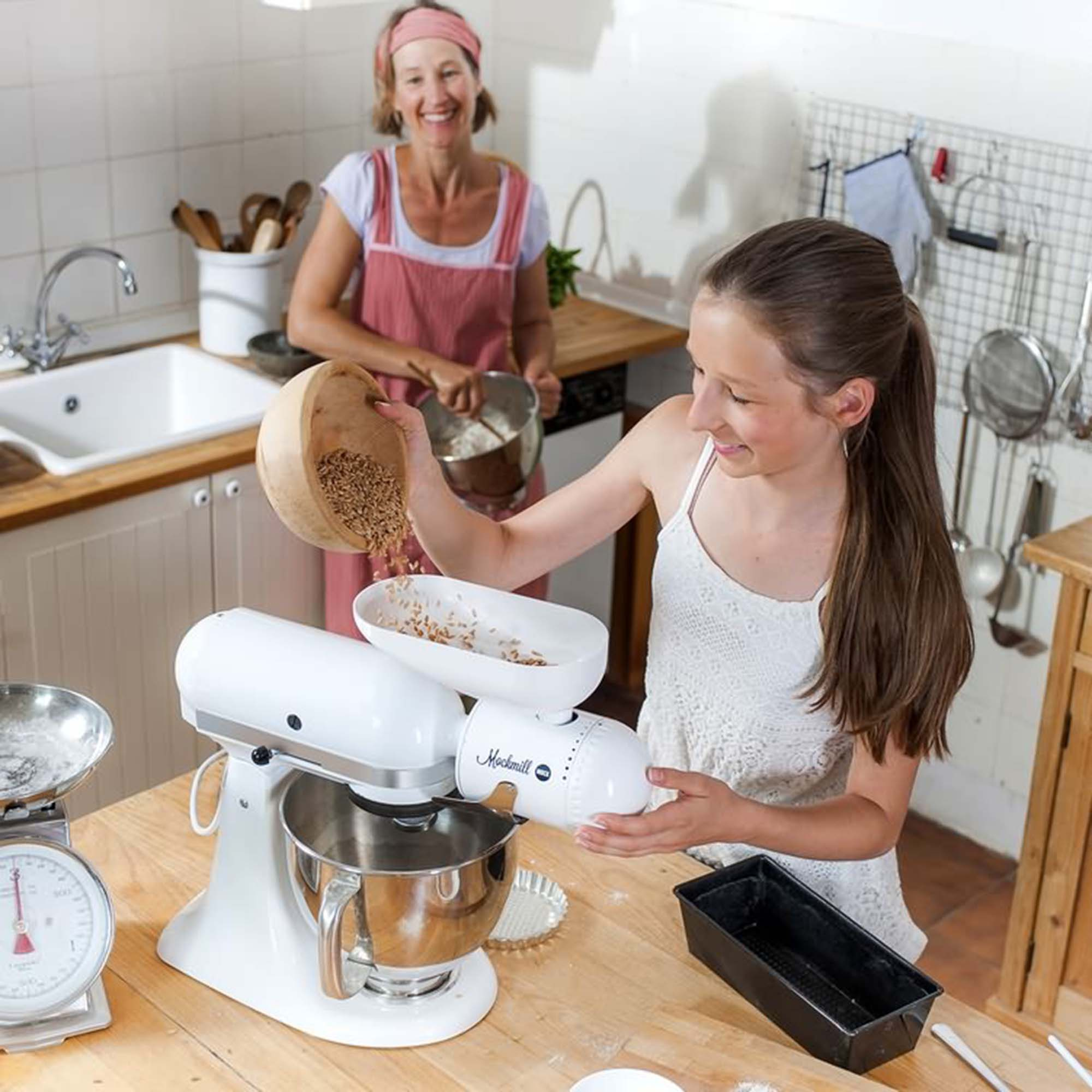 Dziewczyna wsypuje ziarna do miksera. Starsza kobieta stoi w tle, uśmiechając się, w kuchni.