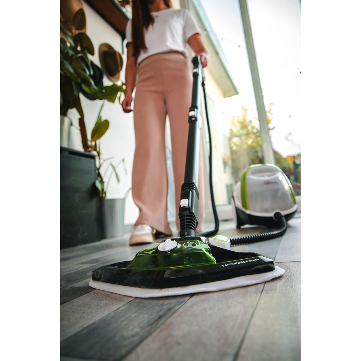Kobieta używająca odkurzacza parowego. Widoczna podłoga i tło. Biało-zielony odkurzacz.