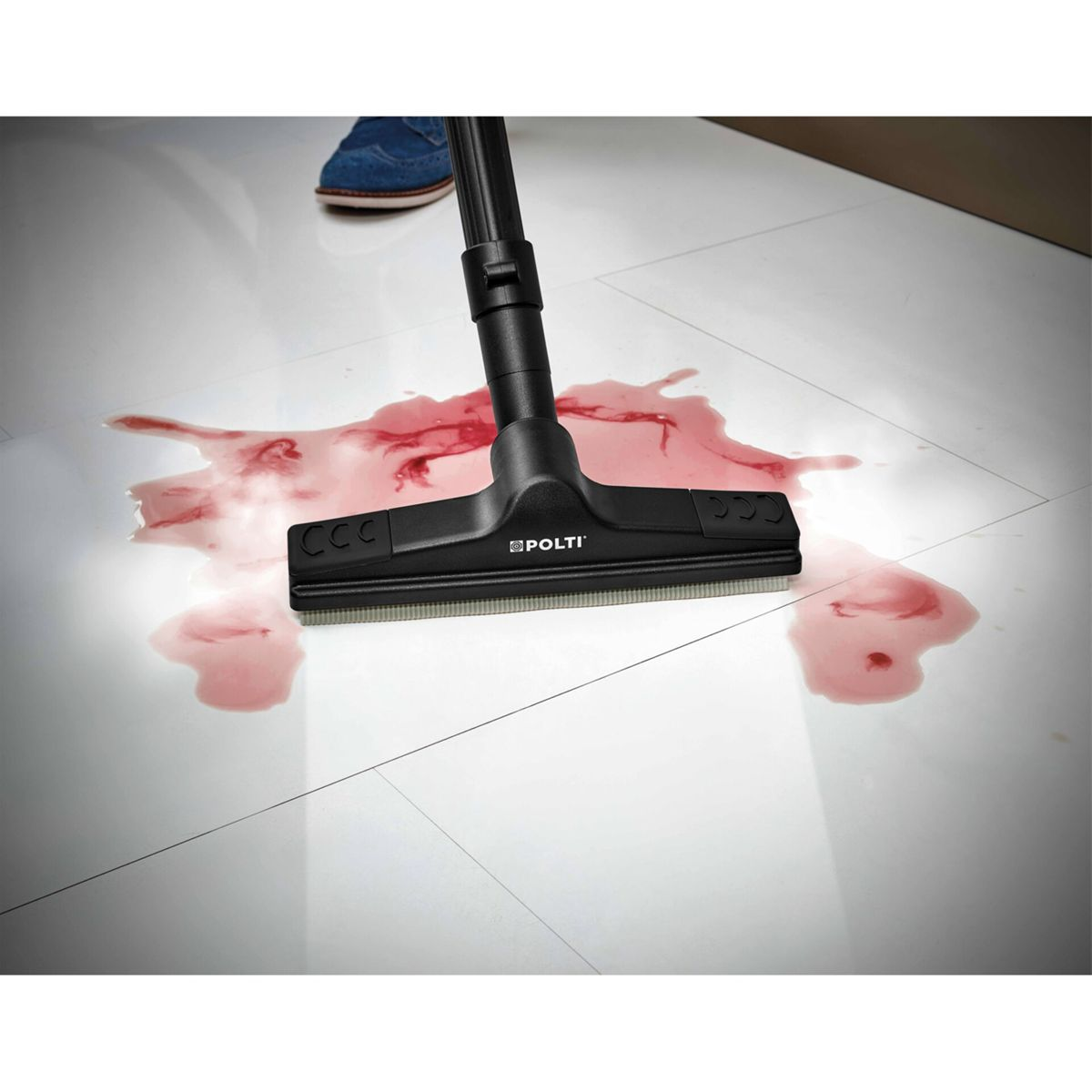 Głowica odkurzacza czyszcząca czerwoną plamę na białej podłodze z płytek. Niebieski but jest w tle.