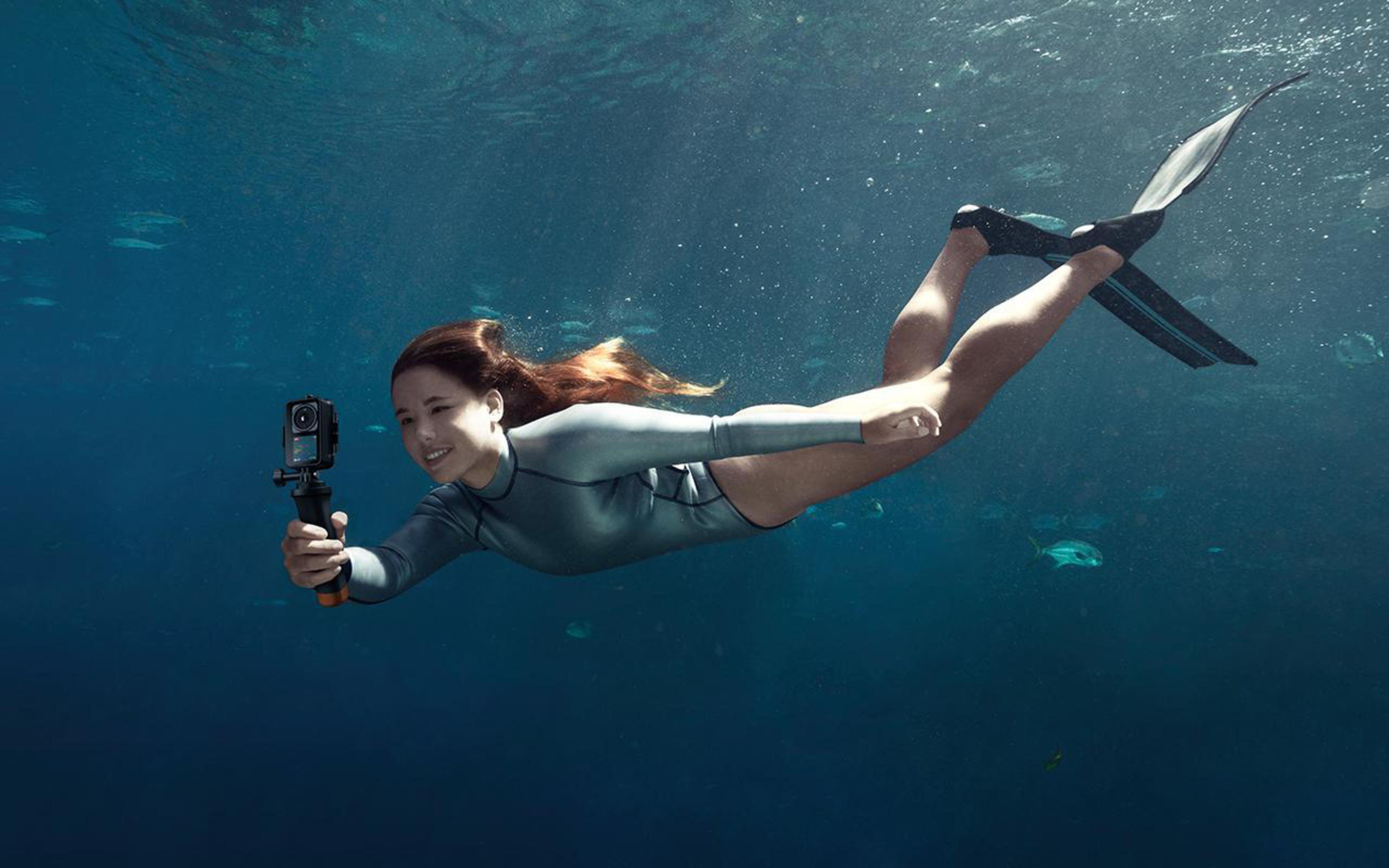 Kobieta pod wodą, trzymająca kamerę. Ma szary kombinezon i płetwy, otoczona rybami.