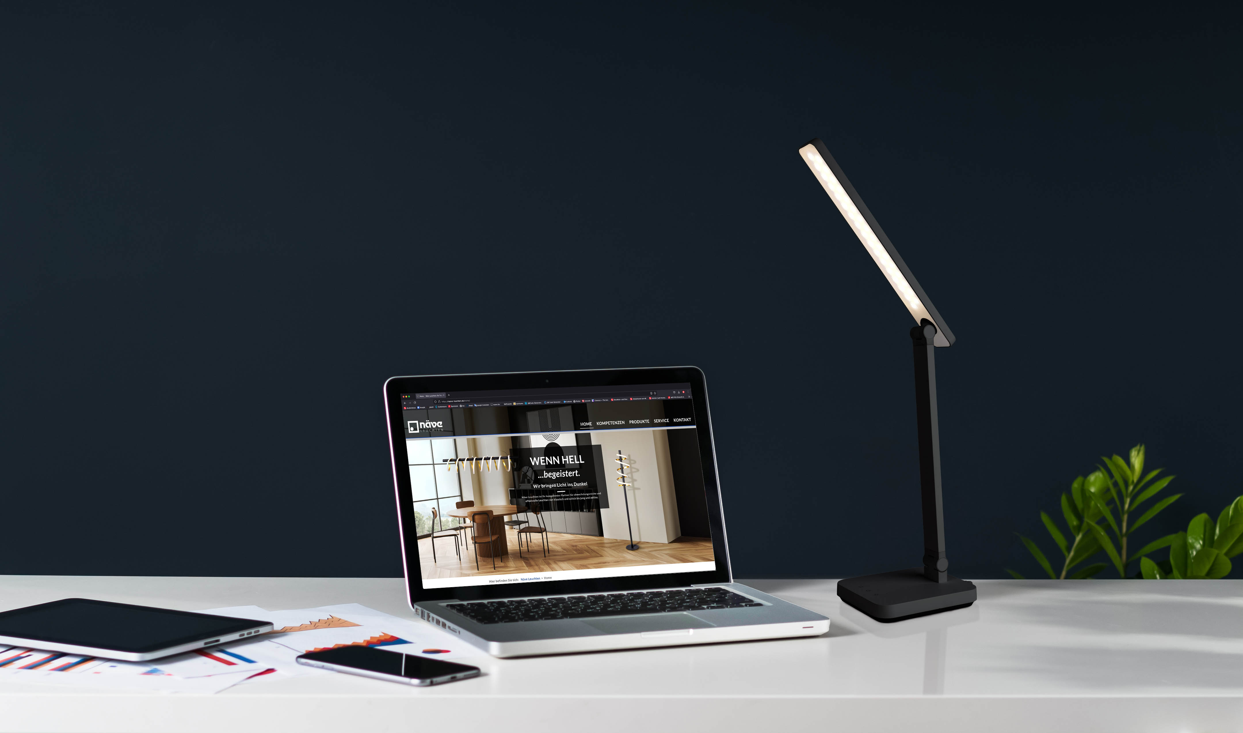Laptop mit Website, Schreibtischlampe, Tablet, Telefon, Dokumente auf weißem Schreibtisch, dunkler Hintergrund.