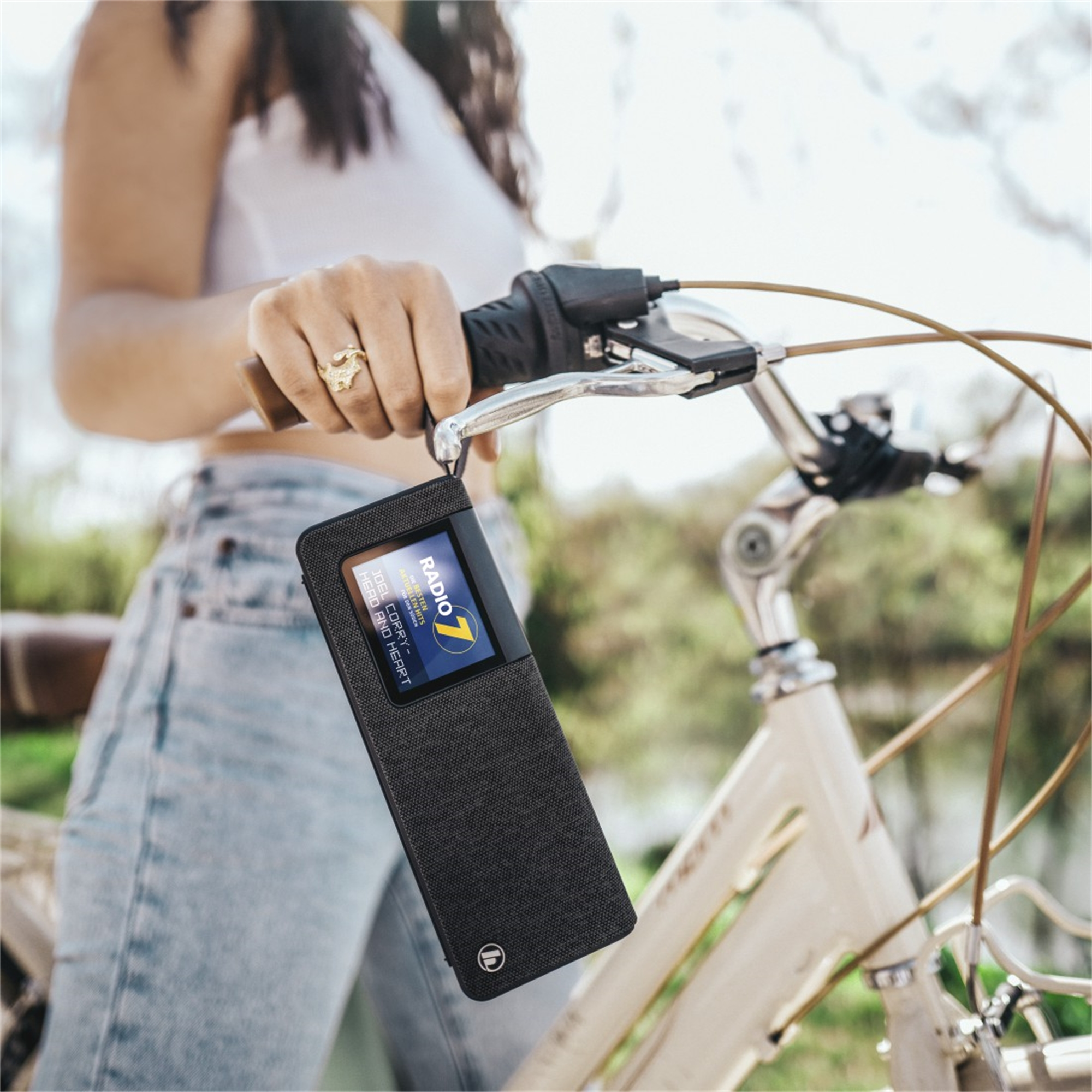 Mujer en bicicleta sosteniendo una radio negra. La pantalla de la radio muestra una emisora. La bicicleta es crema.