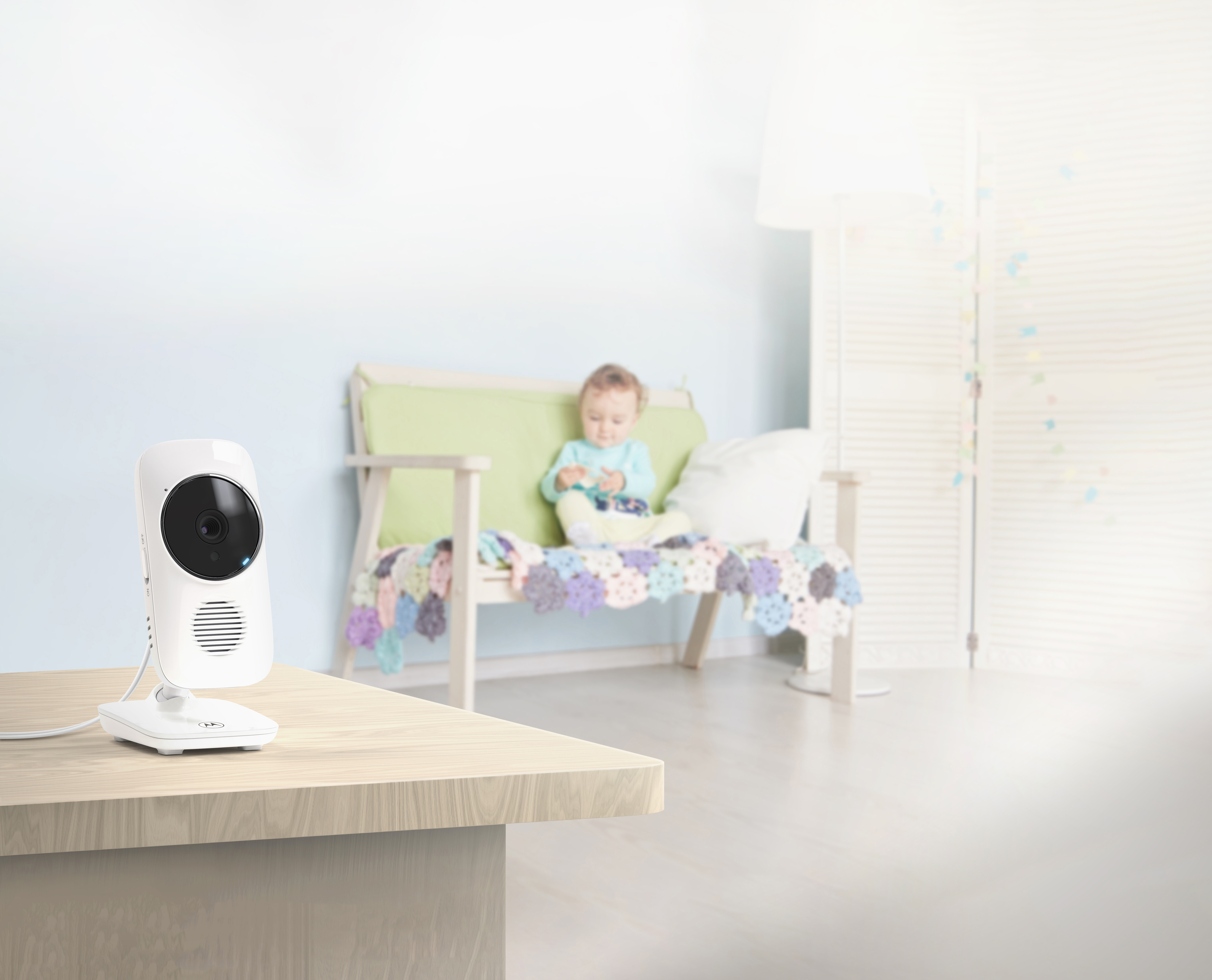 Babyphone auf einem Tisch; ein Baby im Hintergrund auf einer Couch mit Decke und Kissen; heller Raum.