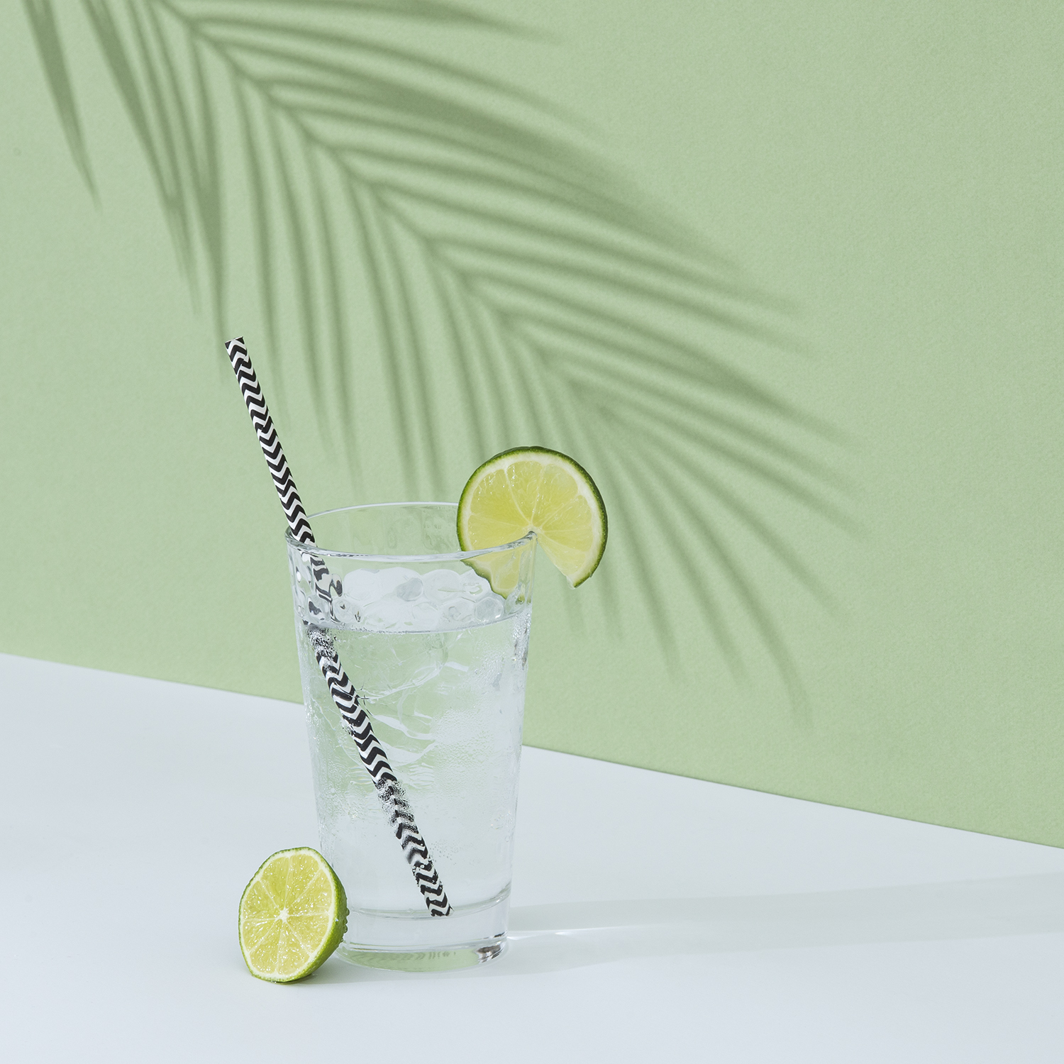 Szklanka wody z lodem, plasterkiem limonki i słomką w paski. Zielone tło z cieniem.