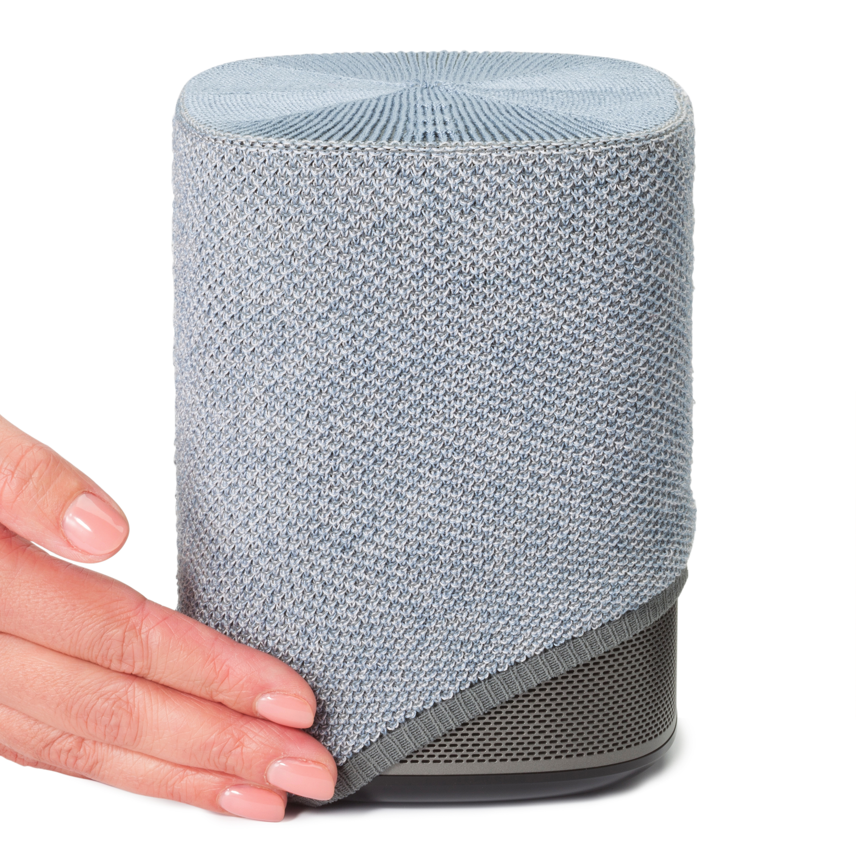 Une main touche un haut-parleur cylindrique recouvert de tissu gris clair, sur fond blanc.