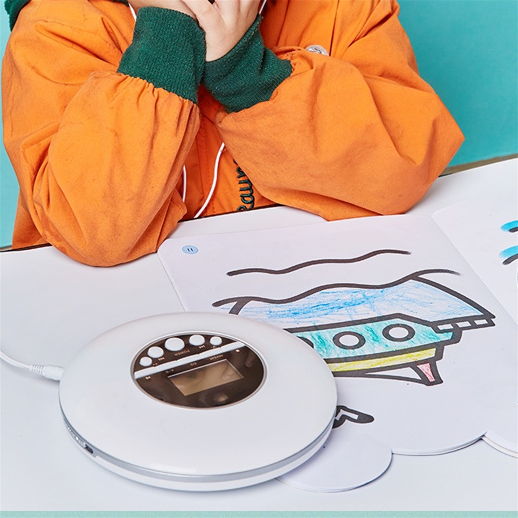Ein Kind in orangefarbenem Hemd zeichnet mit weißem CD-Player und einer Zeichnung einer fliegenden Untertasse.