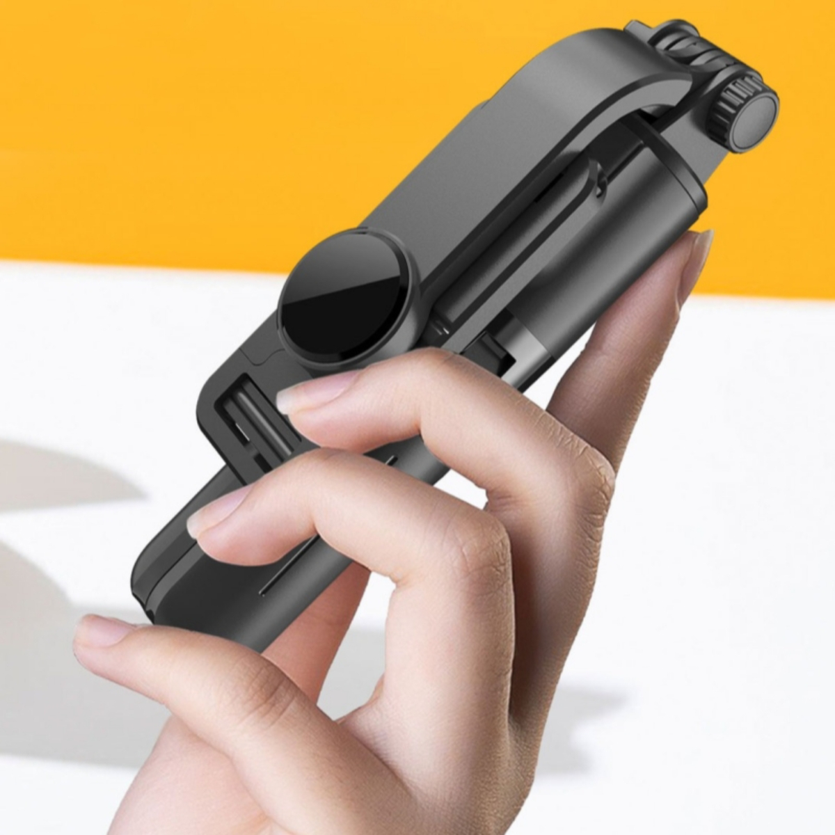 Die Hand einer Person hält einen schwarzen Selfie-Stick, teilweise ausgefahren, auf gelbem Grund.